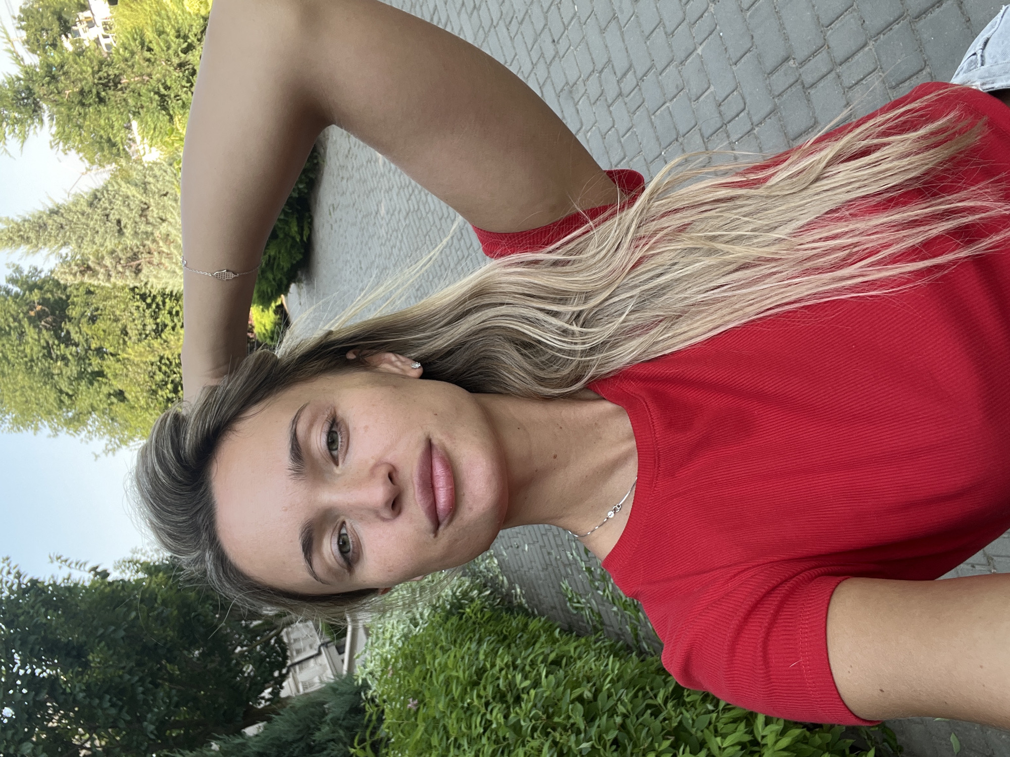 A young woman with long, blonde hair is posing for a selfie outdoors. She is wearing a red t-shirt and a silver necklace. Her right arm is raised behind her head, and she is wearing a bracelet on her right wrist. The background appears to be a park or garden with trees, bushes, and a cobblestone path. The sky is visible in the upper part of the image. The location is identified as Fontanka, Ukraine.