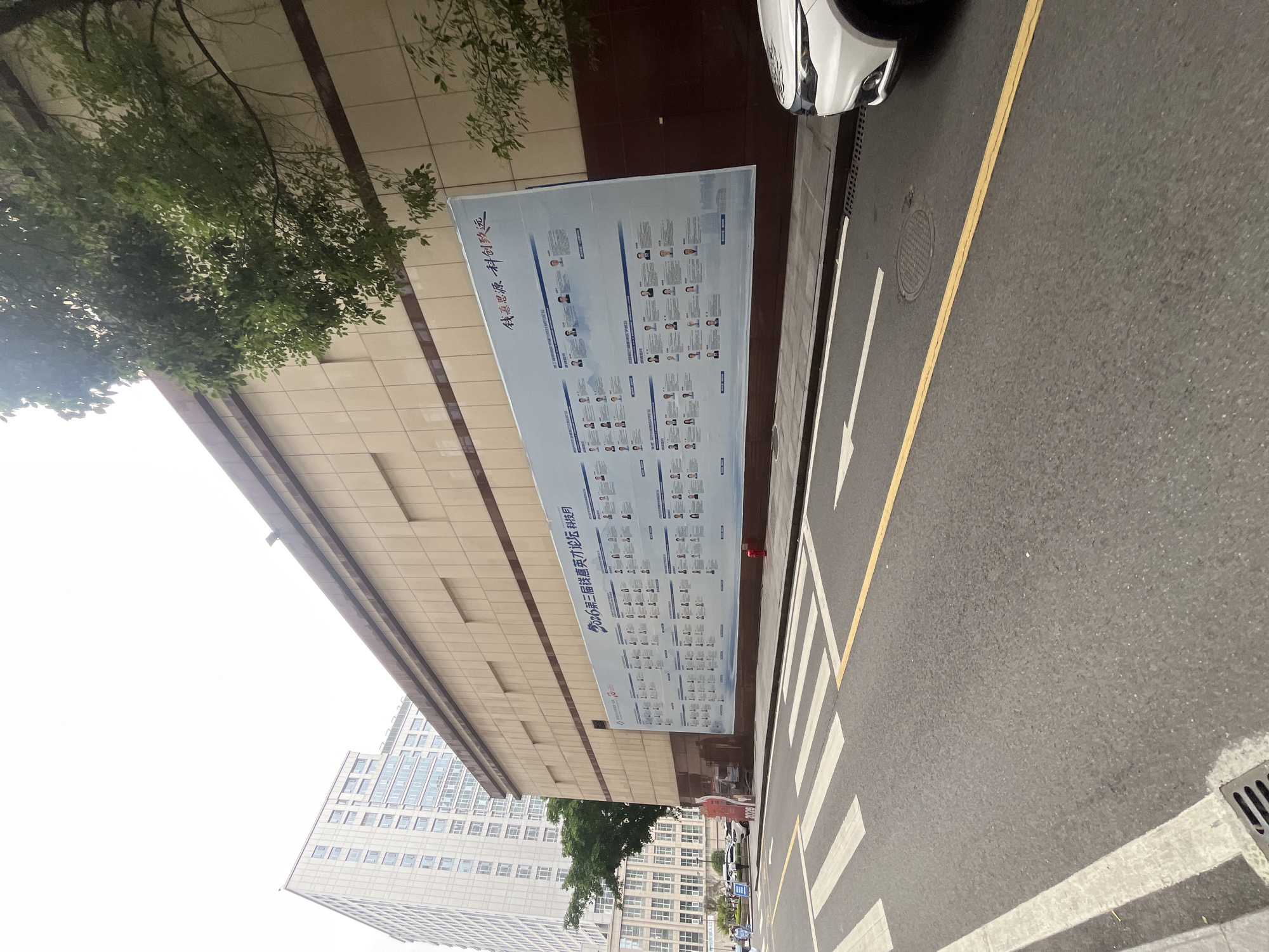 The image depicts a street scene in Yuzhong District, China, with a prominent billboard displayed on the side of a building. The billboard is large and white, featuring multiple rows of individual portraits with accompanying text. The building itself is clad in light-colored square tiles, with darker brown accents along the base and top. A tree with green foliage extends into the frame from the right, partially obscuring the building and billboard.
In the foreground, a pedestrian crossing with white stripes is visible on the asphalt road. A single yellow line runs parallel to the curb. A white vehicle is parked on the right side of the road, with its front partially visible. The sky is overcast and bright. Several people are visible in the distance near the building, some walking and others standing. The overall impression is of an urban environment with a focus on a large informational display.