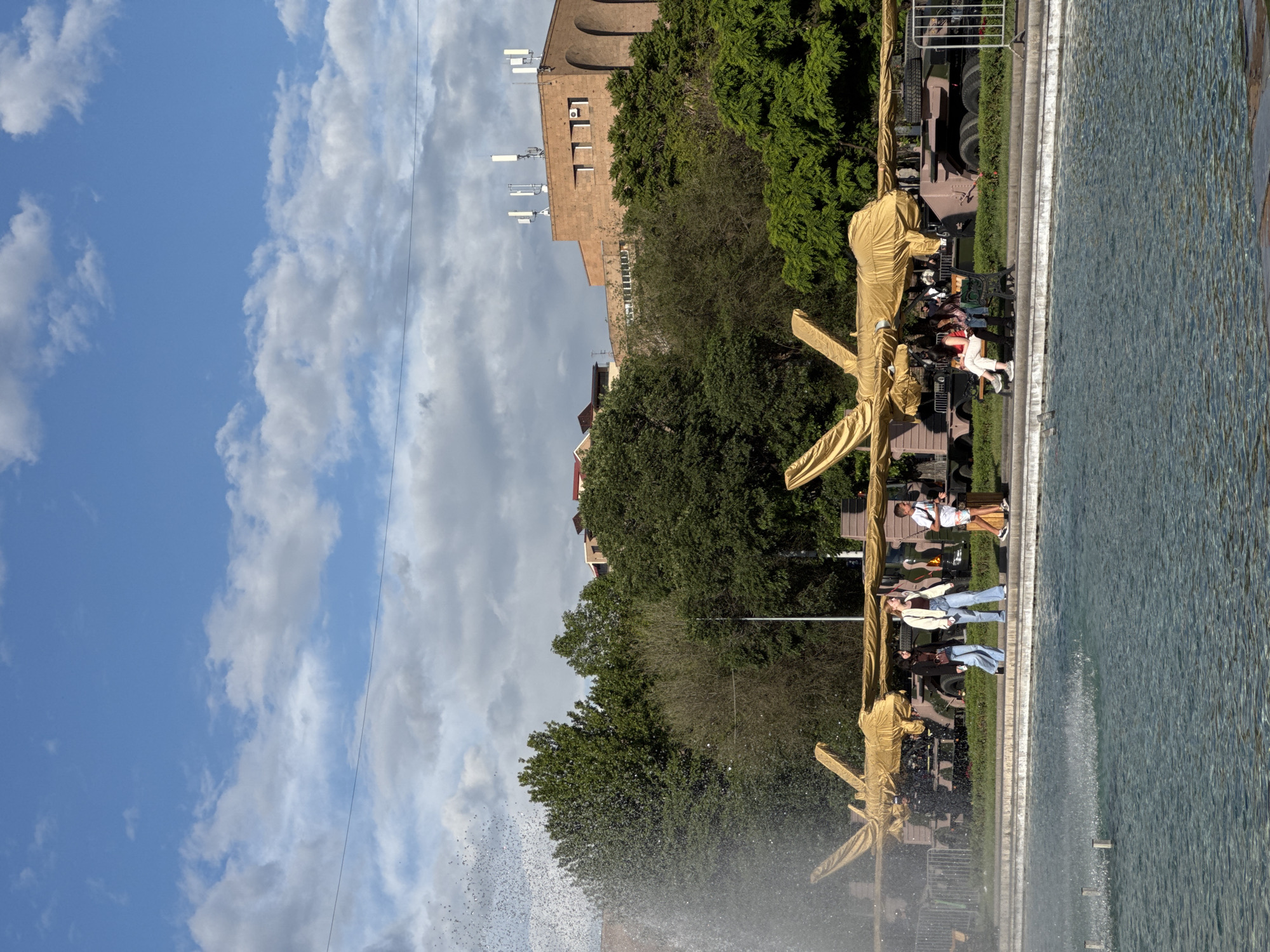 The image shows Yerevan, Armenia, with a fountain in the foreground, its water droplets visible in the spray. Behind the fountain, there are people sitting on benches, some with trucks covered in golden fabric behind them. Trees are situated behind the benches, along with a building further back, topped with antennas. The sky, filled with clouds, is visible above the building.