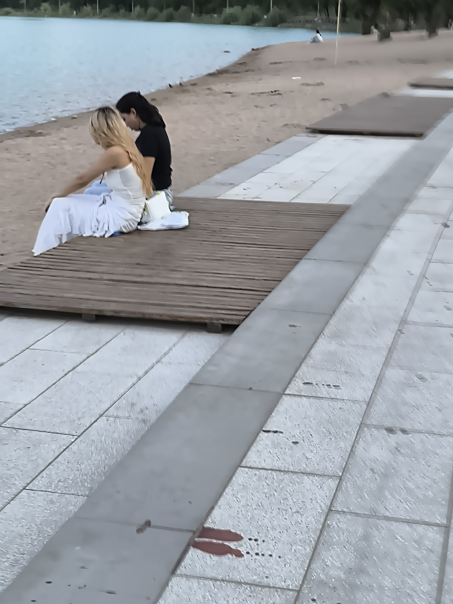 The image shows two individuals sitting on a wooden platform near a sandy beach and a body of water. The person on the left has blonde hair and is wearing a white dress. The person on the right has dark hair and is wearing a black t-shirt. They are positioned back-to-back. The environment suggests an outdoor recreational area, possibly a park or a waterfront promenade. The ground is paved with large, light-colored tiles. In the distance, there is a treeline and the water of a lake or river. The overall scene appears to be a candid moment captured in a public space. The image does not contain any specific landmarks or visual cues that would allow for precise identification of the location within Almaty, Kazakhstan.