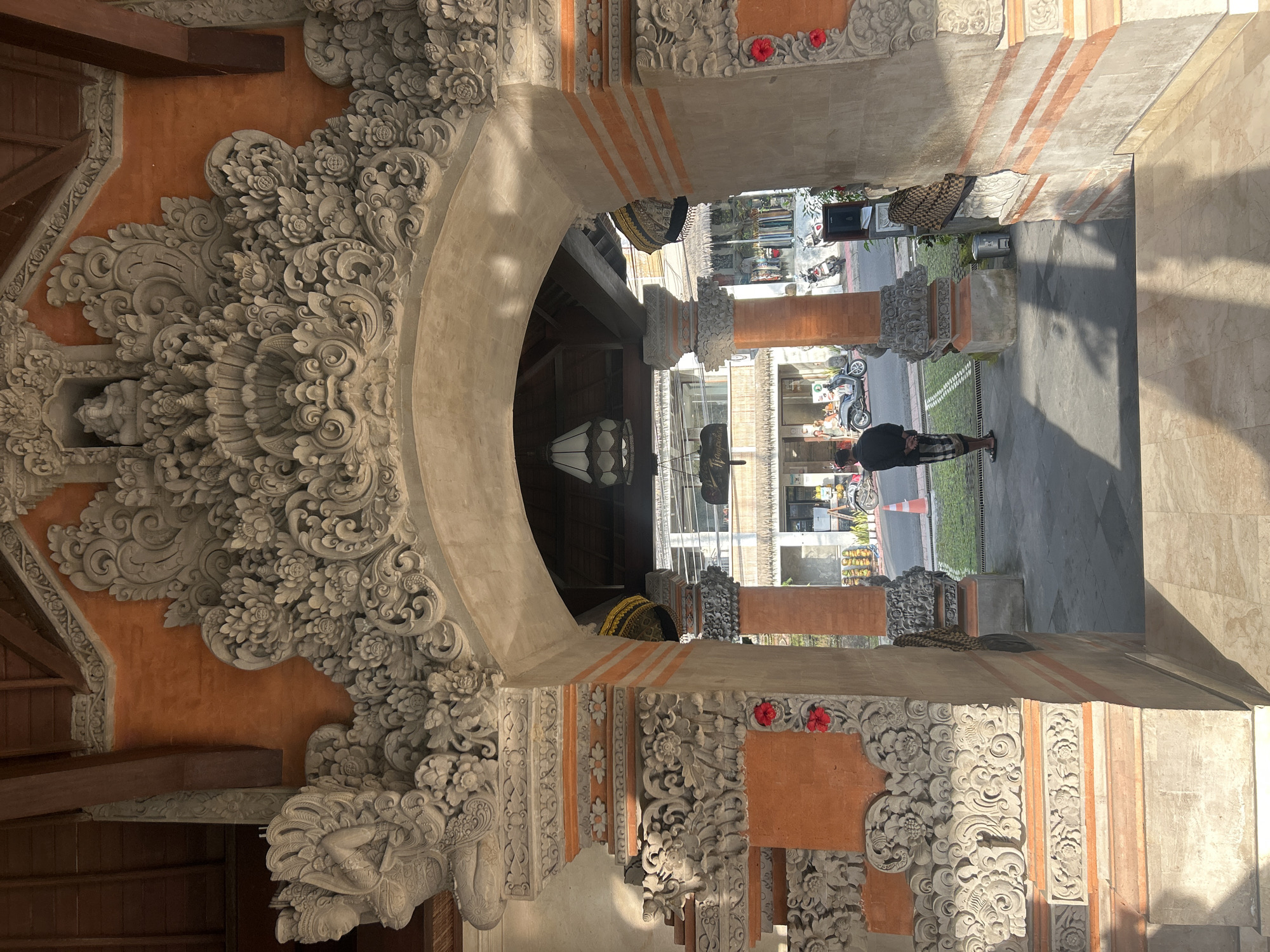 This is a vertical, eye-level, full shot of an intricately carved stone archway entrance to a building in Ubud District, Indonesia. The archway is decorated with detailed floral motifs and stylized figures. Through the arch, a street scene is visible with buildings, parked motorcycles, and a few people. One person, wearing dark clothing and plaid shorts, stands with their back to the camera, facing the street. The lighting suggests it is daytime, with sunlight casting shadows on the ground. The architecture suggests a traditional Balinese style.