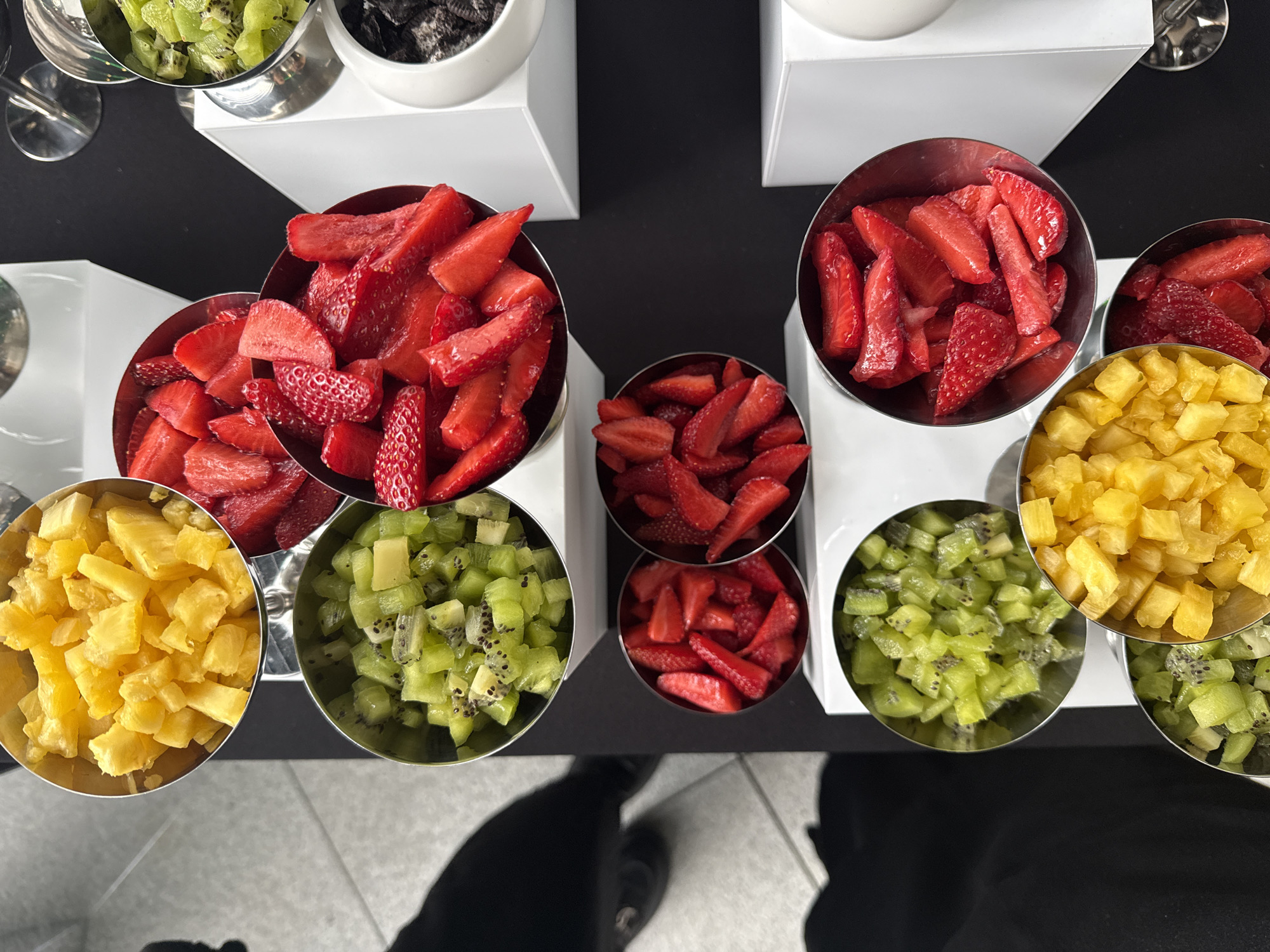 An overhead, slightly tilted view captures a vibrant fruit and topping station set up on a dark surface, likely a black tablecloth or countertop, in Sofia, Bulgaria. The station features numerous stainless steel bowls arranged on white rectangular risers. These bowls are filled with neatly chopped fresh fruits: bright red strawberries, vivid yellow pineapple chunks, and refreshing green kiwi pieces with visible black seeds. The arrangement suggests a self-service buffet or dessert bar.
In addition to the fruits, one white ceramic bowl at the top center holds dark, crushed material, possibly cookie crumbs or a similar topping. Several empty clear glasses are partially visible on the left side of the display, ready for use. Partial views of people's legs, feet, and shadows are visible at the bottom of the image, indicating activity around the station and suggesting individuals are selecting or about to select items. The floor below the table appears to be a light-colored, possibly tiled or concrete, surface. The overall setup is clean, organized, and colorful, offering a healthy and appealing selection for attendees at an event or meal service. No text is visible in the image.