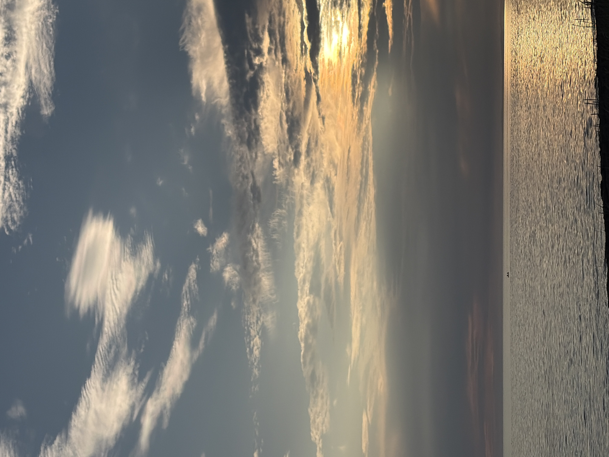 A serene, wide-angle view of a body of water, likely a lake or sea, at sunset. The sky is a canvas of mixed clouds, with soft, wispy white clouds in the upper portion transitioning to darker, more textured clouds lower down. A patch of golden sunlight breaks through the clouds on the right side, reflecting a warm glow onto the water. The water itself is calm, with gentle ripples creating a textured surface. A faint golden shimmer of sunlight dances across the water’s surface. In the distance, a small, dark silhouette of what appears to be a boat with at least two occupants is visible on the water. The shoreline in the foreground is dimly visible, with indistinct dark shapes suggesting vegetation or structures. The overall impression is one of peaceful twilight in a natural setting, possibly near Ozerskoe, Russia, given the context.