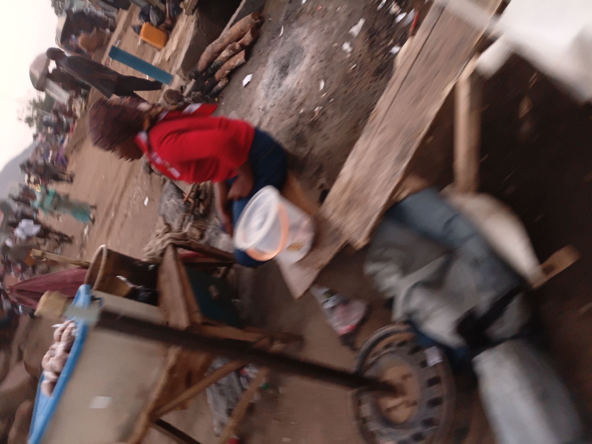 The image depicts an outdoor market scene in Gwong, Nigeria. A person wearing a red top and dark trousers is seated on a wooden plank. In the foreground, a blue container filled with what appears to be meat is visible, resting on a wooden stand. Various objects, including buckets and debris, are scattered around the ground. The background shows a bustling market with several people and structures, suggesting a lively commercial environment. The overall impression is one of a typical market day in an urban African setting.