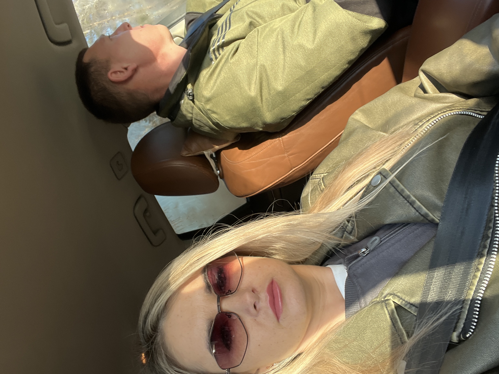 A woman with blonde hair and sunglasses is taking a selfie inside a car. She is wearing a green jacket and has a seatbelt on. Behind her, a man with short dark hair is reclined in the passenger seat, also wearing a green jacket. The car interior is visible, including brown leather seats and parts of the car's ceiling and window. There is no information about location in the city of Chaikovsky, Russia.