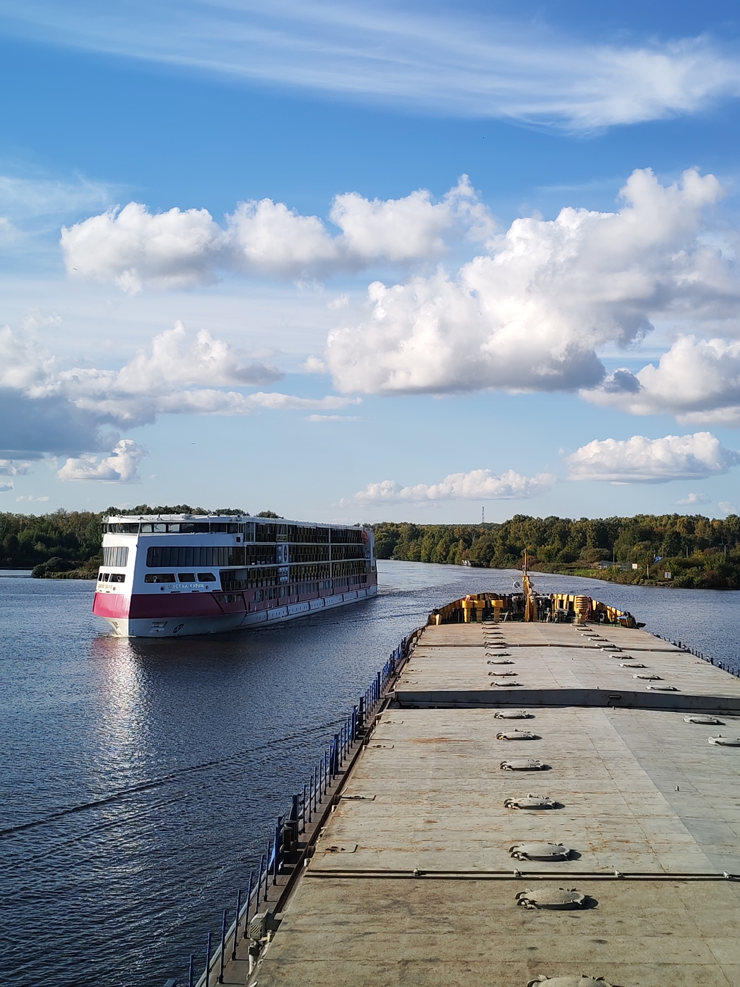 The image captures a wide, calm river under a bright blue sky with scattered white clouds. On the left side of the river, a large, modern passenger ship with a distinctive pink and white hull is sailing. The ship appears to be a multi-deck vessel, with numerous windows visible along its sides. On the right side of the river, the viewpoint is from the deck of a long, flat barge, also traveling on the water. The barge's deck is visible in the foreground and is covered with a patterned concrete surface and has numerous circular hatches. The riverbanks on both sides are lined with dense, green trees, suggesting a natural, undeveloped area. The overall scene depicts a serene day on the water, with two vessels navigating the waterway. The specific location within Drachevo, Russia, cannot be determined from the visual information provided in the image.