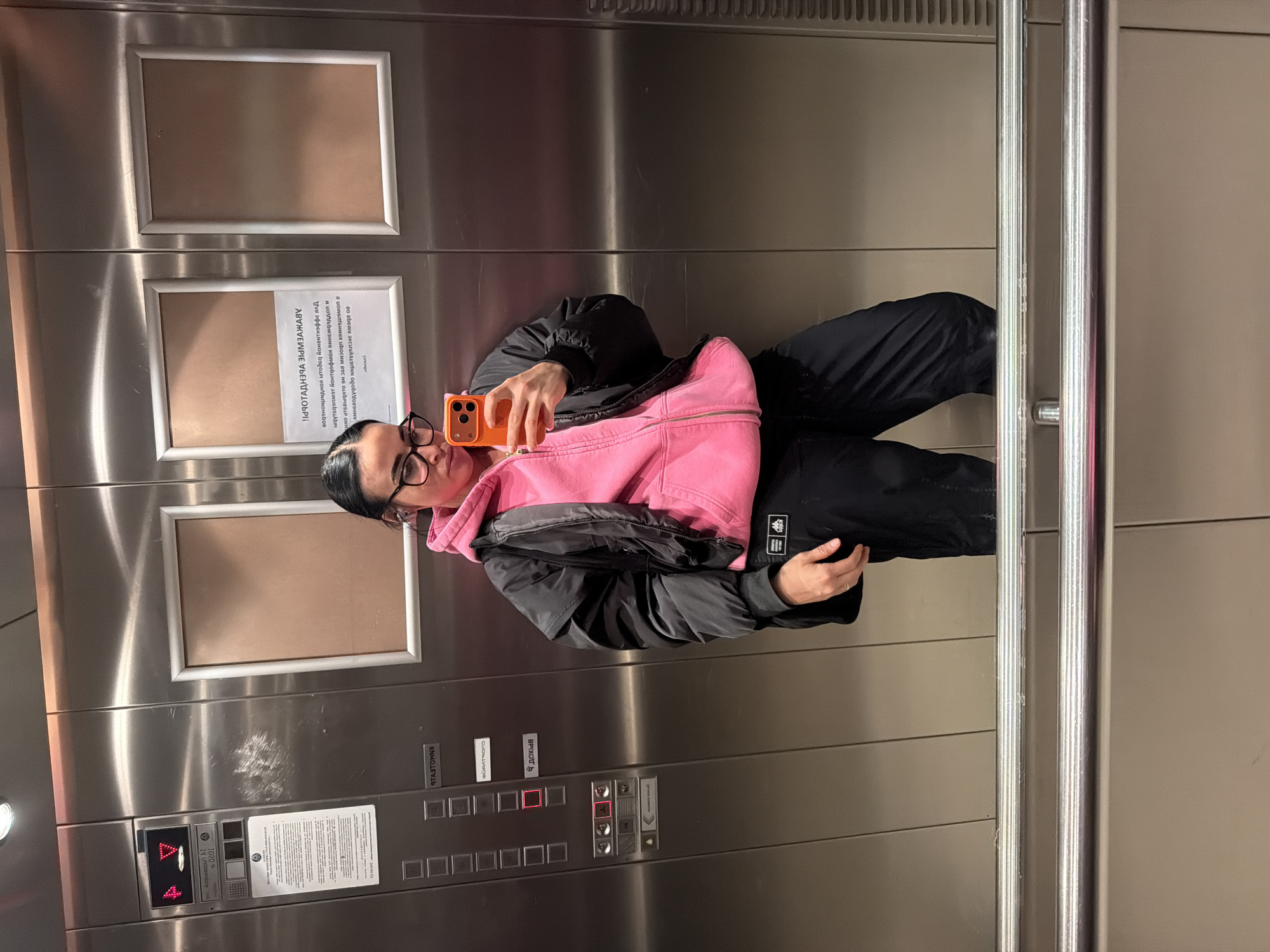 A woman in a pink sweatshirt and black jacket stands in an elevator taking a selfie with an orange phone. The elevator appears to be in Russia, given the Cyrillic text on the elevator control panel and signs. The city cannot be determined. The elevator has a metallic interior with two framed areas on the back wall, one with a paper notice. The woman is wearing glasses and her hair is pulled back. The elevator doors have metal handles.