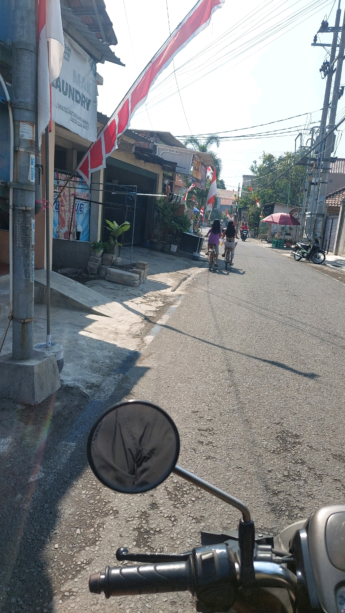 The image shows a street scene in Sidoarjo, Indonesia, on a sunny day. The viewpoint is from the perspective of a motorcycle rider, as indicated by the presence of the handlebars and a rearview mirror in the foreground. The street is paved and lined with buildings on either side.
On the left side of the street, there is a building with a sign that says "MAK LAUNDRY". There are also Indonesian flags displayed, with red and white stripes, one of which is a long banner draped across the street. A concrete pole with various signs is also visible.
On the right side of the street, there are more buildings, some with awnings and small shops. A red umbrella is set up, suggesting a street vendor or seating area. Several Indonesian flags are also visible on this side.
In the middle of the street, two individuals on bicycles are riding away from the camera. The person in front is wearing a purple shirt and the person behind is wearing a patterned top. A motorcycle is parked on the right side of the street. The sky is clear and bright blue, with power lines visible against it. The overall impression is a typical urban street in Indonesia, likely decorated for a national celebration or event given the prominent display of flags.