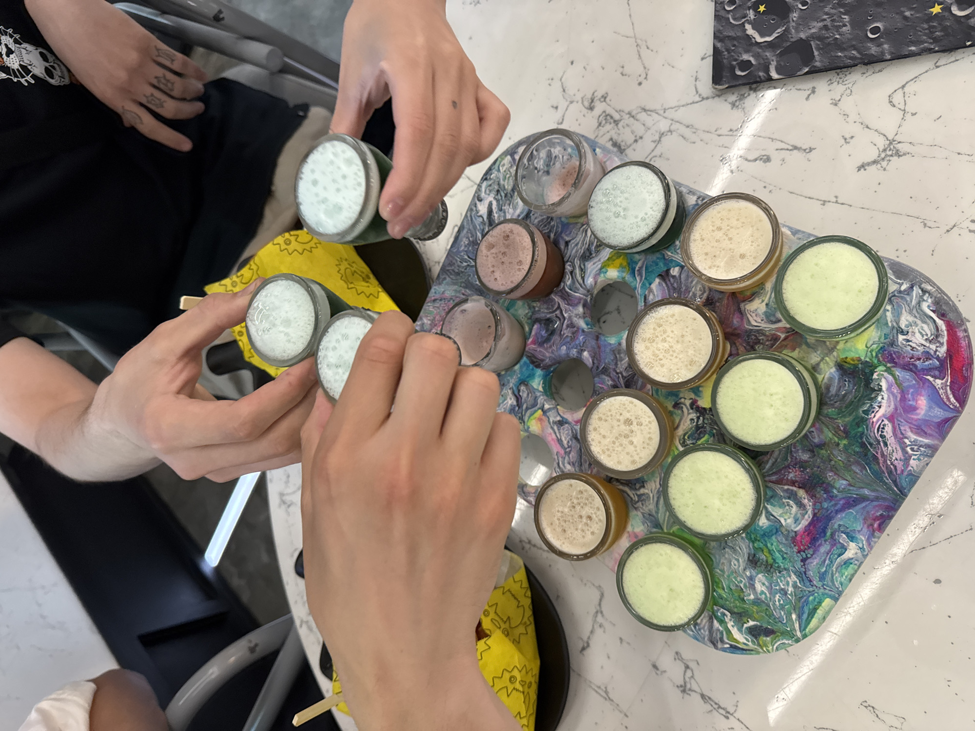 The image shows a top-down view of a table with several small glasses filled with colorful drinks. Three people's hands are visible, interacting with the glasses. One person is holding a glass with a white, frothy liquid, and another is reaching for a glass from a multi-colored tray. The tray itself is decorated with a swirling, abstract pattern of purples, blues, greens, and white, resembling a pour painting.
The drinks in the glasses vary in color, including white, light green, pale yellow, and a light pinkish-brown. Some glasses contain bubbly liquids. The table surface has a white, marble-like pattern. In the background, a black t-shirt with a white graphic of a skull is partially visible. A yellow napkin with a black zig-zag pattern is also present. To the upper right, a decorative item with a moon and stars motif, possibly a placemat or coaster, is visible on the table.
The scene appears to be set indoors, possibly in a cafe or a casual gathering. The lighting suggests it could be daytime. The hands and the way they are reaching for the drinks suggest an activity like a tasting or sampling of beverages. There are no clear emotional cues, but the presence of multiple people and drinks implies a social interaction. No visible text is present in the image.