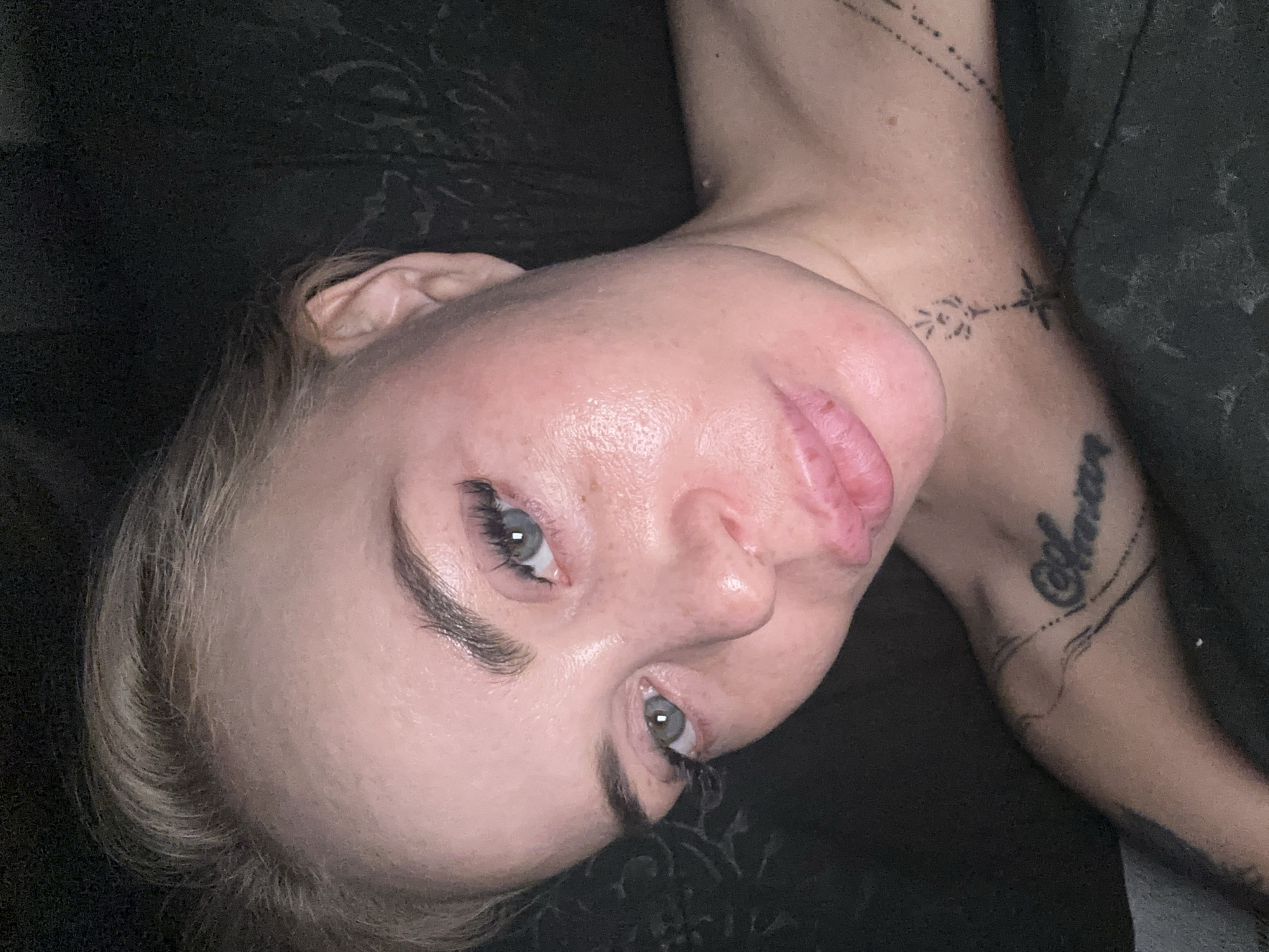 An extreme close-up selfie of a woman's face and upper chest is captured in a dark environment. The woman has fair skin with some redness and a slightly oily sheen on her forehead and cheeks. Her blonde hair is pulled back, revealing her forehead and ear. She has light blue eyes, dark eyebrows, and is wearing false eyelashes. Her lips are full and slightly parted. On her left shoulder, a tattoo is visible with the word "Amor" in script. Further down her neck and chest, there are two delicate line tattoos: one resembling a flower with radiating lines, and another of a dotted line with small dashes. The background is dark and appears to be a textured surface. The woman is lying down, and the lighting is dim, highlighting her facial features.