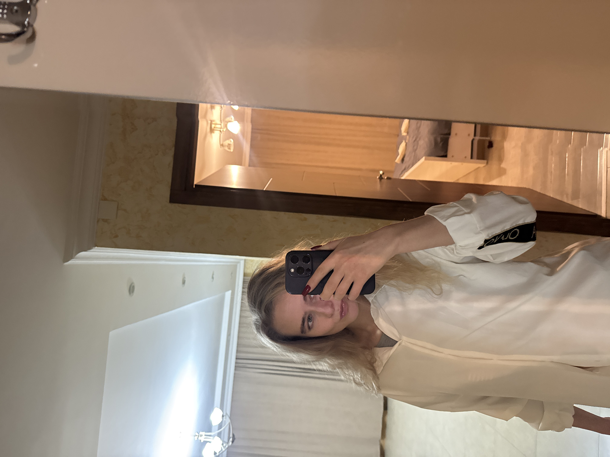 A woman is taking a selfie in front of a mirror. The location appears to be a residential bathroom with cream-colored walls, white trim, and recessed lighting. The mirror reflects the bathroom, which includes a vanity with lighting, a bed and chair, and a window covered by a curtain. The woman is wearing a white jacket with black-and-white lettering on the sleeve and holding a smartphone. The image appears to be rotated clockwise.