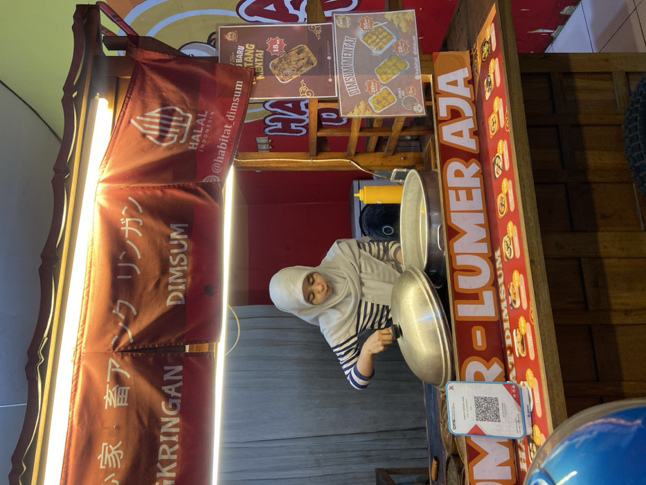 A female vendor operates a dimsum food stall in Tenggulunan, Indonesia. The woman, wearing a light grey hijab and a blue-and-white striped long-sleeved shirt, is focused on preparing food within a metallic steaming pot. She holds the lid of a large, domed steamer, which rests partially open beside another open steaming pot on the wooden counter. To her right, a yellow squeeze bottle and a small black fan are present.
Above the stall, illuminated by overhead fluorescent lights, hang maroon banners with white lettering displaying "KRINGAN DIMSUM," Japanese characters, and a "HALAL INDONESIA" logo with the social media handle "@habitat_dimsum." The front of the wooden counter features an orange sign with the text "LUMER AJA," below which is a red strip illustrating various dimsum options. A QRIS payment stand with a QR code is visible on the counter.
To the right of the vendor, two hanging advertisements are displayed: a brown poster for "MENU BARU TANG MENTAI" priced at 18,000, and a yellow poster showcasing "DIMSUM MENTAI" with options priced at 20,000 and 35,000. The stall's immediate background is a plain grey, transitioning to a vibrant orange and yellow wall on the far right. A blue object, possibly a helmet, is partially visible in the bottom-left foreground.