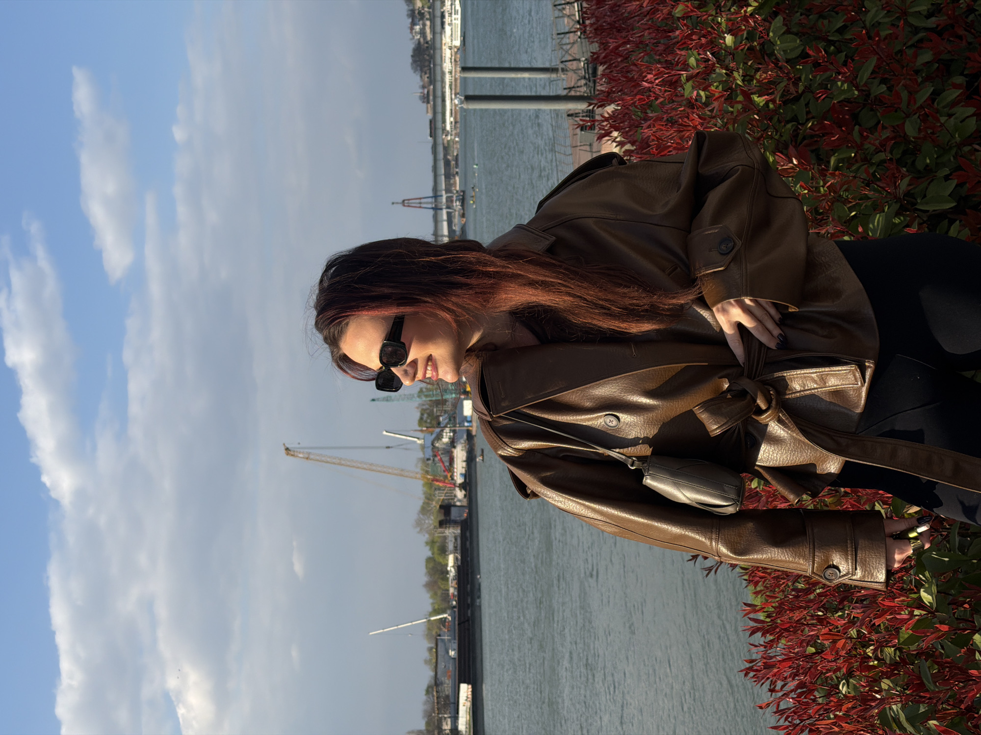 A female subject with long, reddish-brown hair and dark rectangular sunglasses stands outdoors, with her head turned to the right, exhibiting a slight smile. She wears a brown, belted trench coat over dark clothing and a small, grey, quilted crossbody bag. Her left hand holds a thin, cylindrical object with yellow accents, possibly a vape pen. In the immediate foreground, dense bushes with prominent red-tipped foliage are visible. The background features a wide river (the Sava River in Belgrade, Serbia), with a significant construction site on the far bank, characterized by multiple large cranes and industrial structures. Further downstream, the distinctive red and blue pylons of the Ada Bridge are visible, with distant urban elements and hills beyond. The scene is illuminated by natural light under a partly cloudy sky. The location is consistent with the Belgrade Waterfront area along the Sava River, looking towards the confluence with the Danube and the old city.