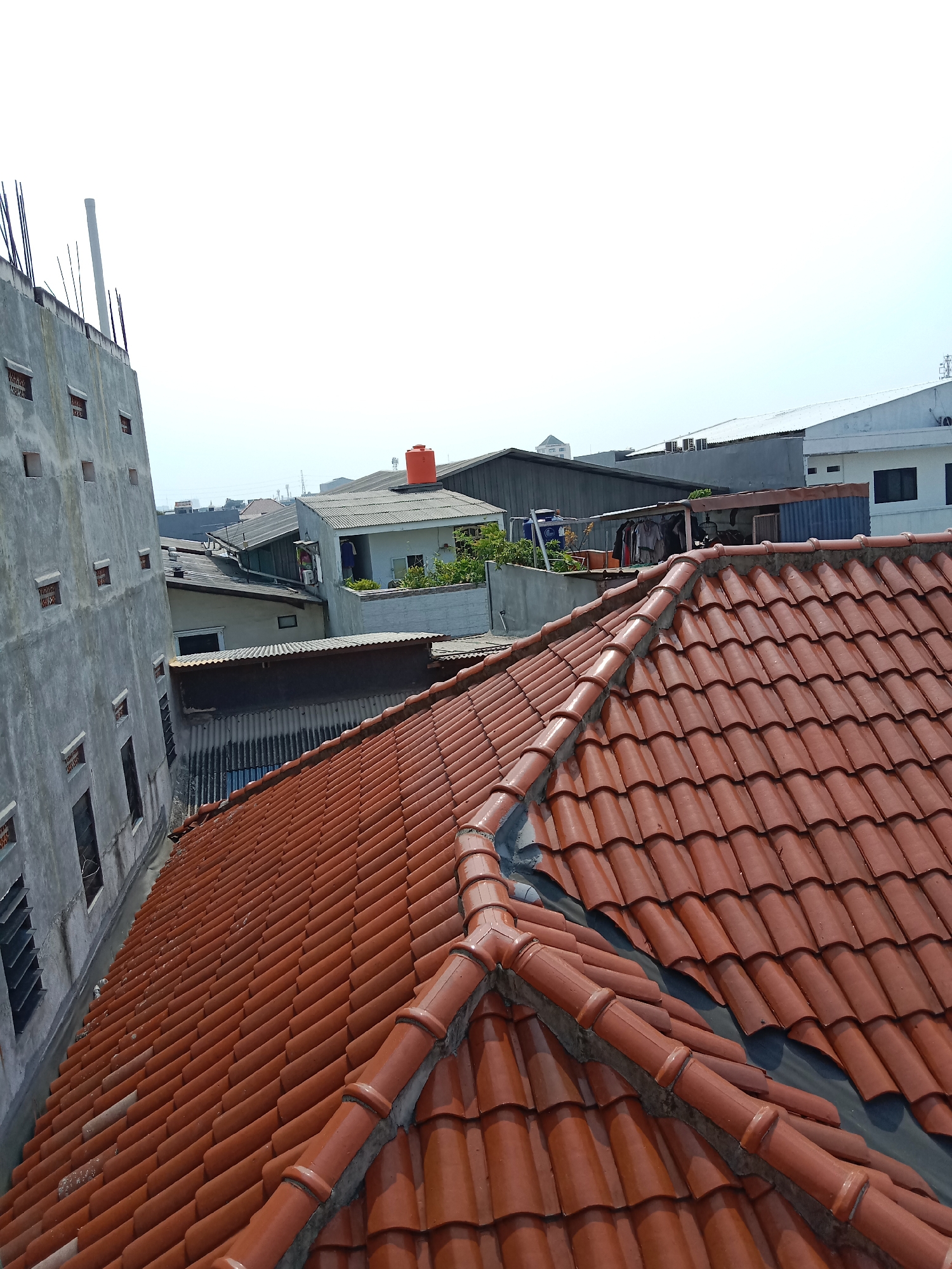 The image provides an elevated view of a dense urban residential area, likely in West Jakarta, Indonesia, characterized by numerous rooftops and buildings. In the foreground, a prominent reddish-brown tiled roof dominates the right side of the frame, with visible curved tiles and a ridge detail. To the left, a tall, grey concrete building with visible windows and ventilation openings suggests construction or renovation, with rebar protruding from its top.
Beyond these immediate structures, a collection of other buildings with various roofing materials, including corrugated metal and tiles, are visible. Some structures appear to be houses, while others may be workshops or small commercial buildings. A bright orange water tank is perched atop one of the buildings further in the background, a common sight in Indonesian urban landscapes. The overall environment appears to be a densely populated neighborhood with limited green space. The sky is bright and overcast, indicating daylight conditions, and there are no people clearly visible in the scene.