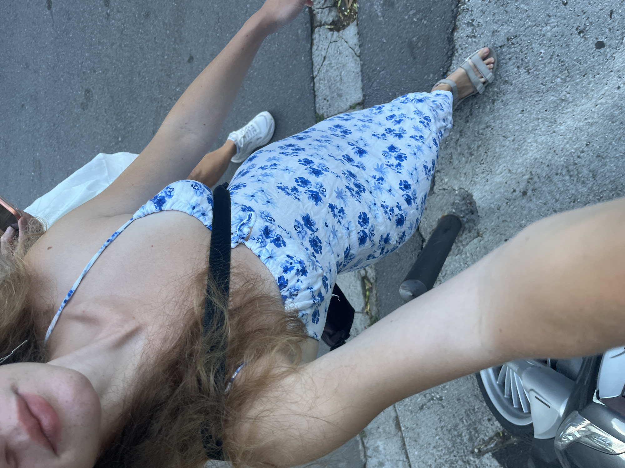A young woman stands on a paved surface, likely a street in Budva, Montenegro, given the presence of a moped in the foreground. She is wearing a white and blue floral print dress, a black strap across her chest, and grey sandals. Her hair is long and light brown. The background consists of a grey asphalt road and a concrete sidewalk. Her left leg is bent, and her right foot is positioned slightly forward. A white plastic bag is partially visible behind her.