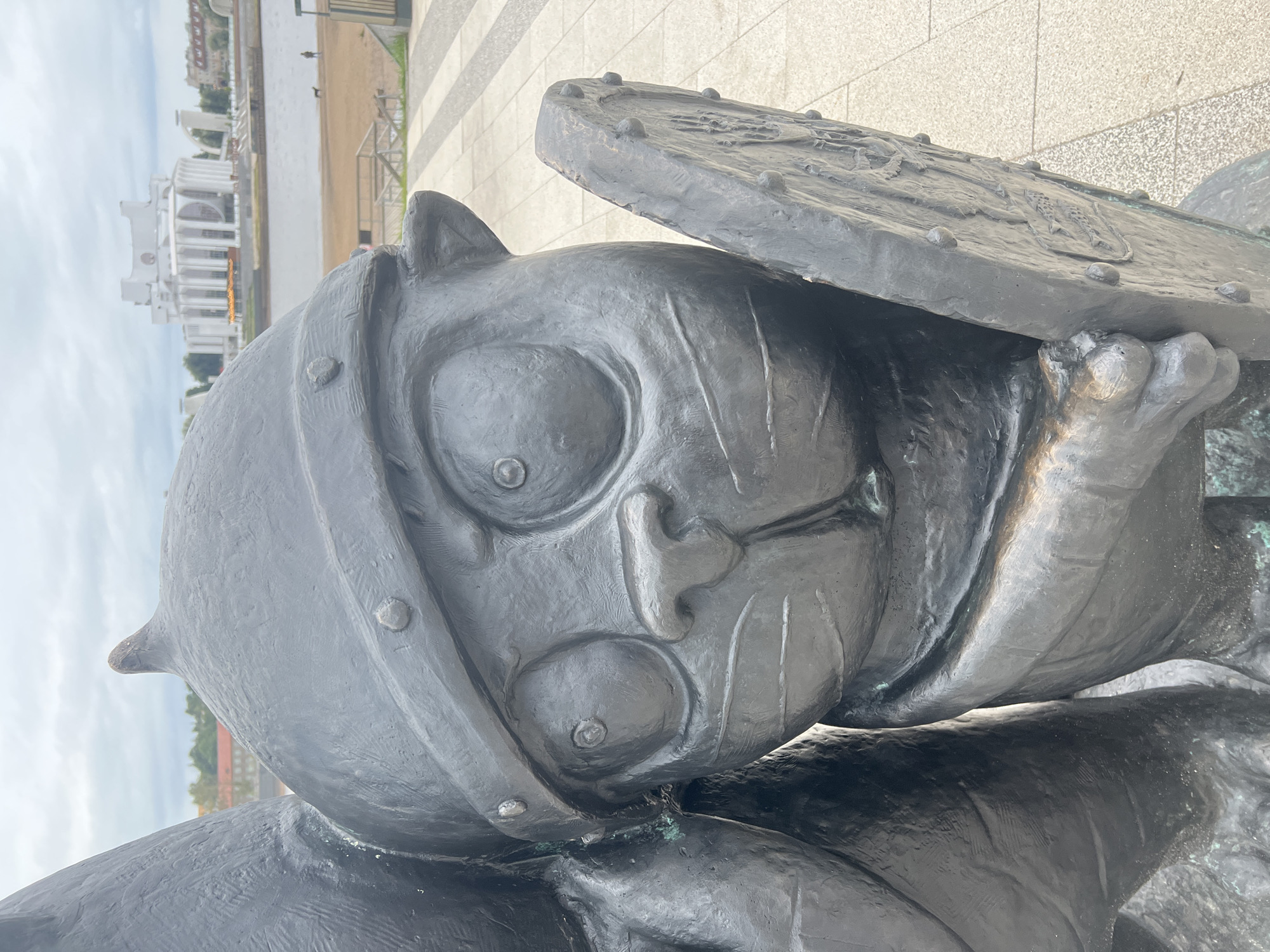 This is a medium-shot, eye-level, outdoor photograph of a bronze sculpture of a cartoonish animal wearing a helmet and holding a shield. The sculpture is dark, with a rough texture and some green patina. The animal has large, round eyes, a small nose, and a mouth with visible lines. It appears to be a cat or a similar feline creature. The helmet is round with a pointed top and decorative studs. The shield is also round and appears to have some embossed details.
Behind the sculpture, a body of water is visible, and across the water, a large, white, modern building with columns stands out against a cloudy sky. To the right of the water, there is a paved area with a sandy beach beyond. A few people and a dog are visible in the distance on the beach. The ground in front of the sculpture is paved with light-colored tiles in a geometric pattern.
This sculpture is located on the embankment of the city of Veliky Novgorod, Russia, along the Volkhov River. It is part of a series of whimsical sculptures dedicated to the heroes and motifs of Russian folklore, specifically the animated film "The Snow Queen" by the Soyuzmultfilm studio. This particular sculpture represents the character of the Snow Queen's messenger, a talking cat.