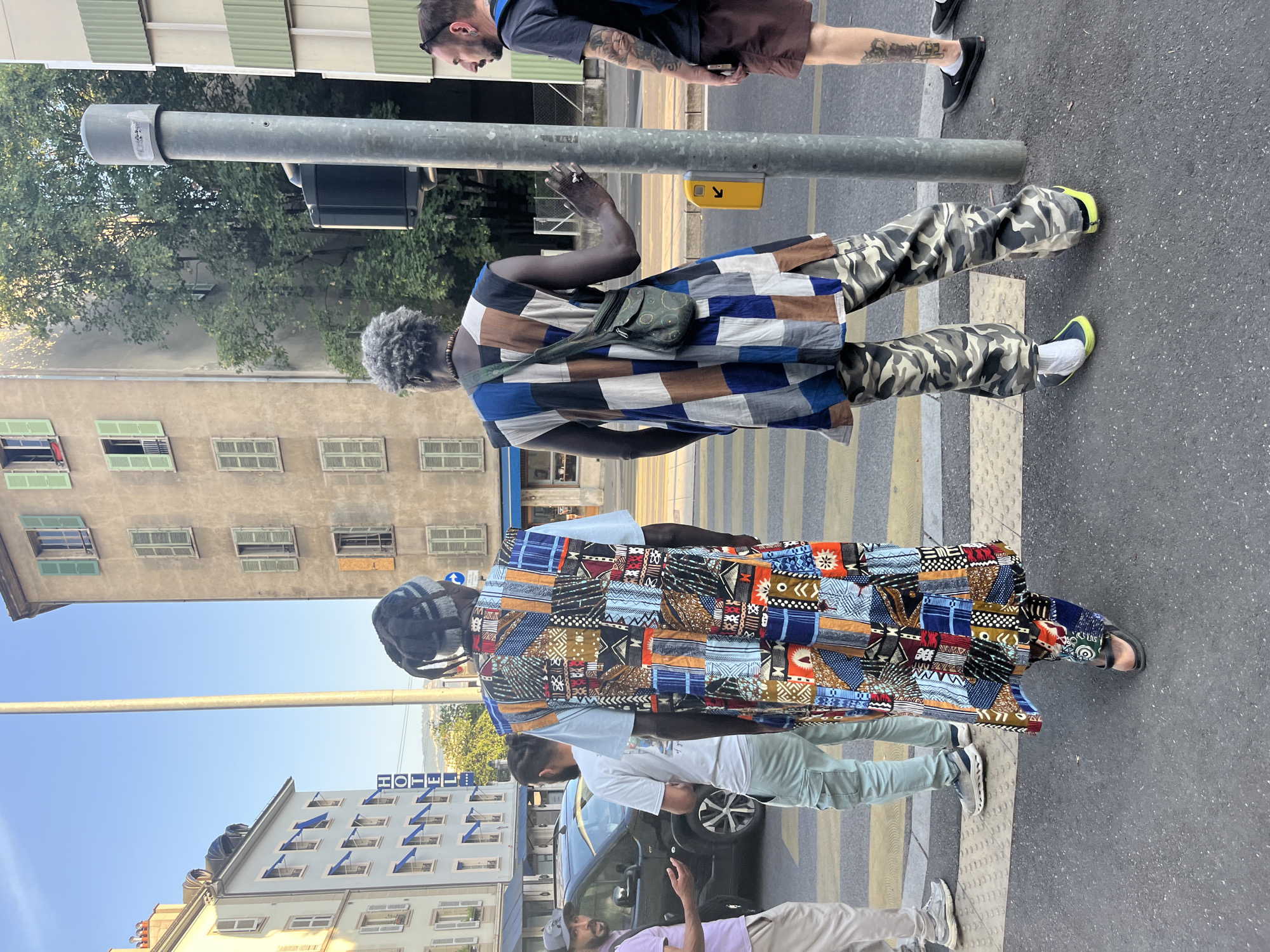 The image shows a street scene in Geneva, Switzerland, likely on a sunny day given the lighting. In the foreground, two men are standing on a pedestrian crossing. The man on the left is wearing a patterned long vest over a light blue t-shirt and light-colored cargo pants. The man on the right is wearing a colorful, patchwork sleeveless garment with a brown and blue checkered pattern, and camouflage pants. He has a small green bag slung across his chest. A third man with tattoos on his arm is visible on the right, wearing a dark t-shirt and brown shorts.
Behind the men, there are buildings with typical European architecture. One building on the left has a sign that reads "HOTEL." There is a black car stopped at the intersection, and other people are visible in the background. A traffic light pole stands to the right of the main subjects. The pedestrian crossing is marked with yellow and white lines. The environment suggests an urban setting with clear visibility and good weather.