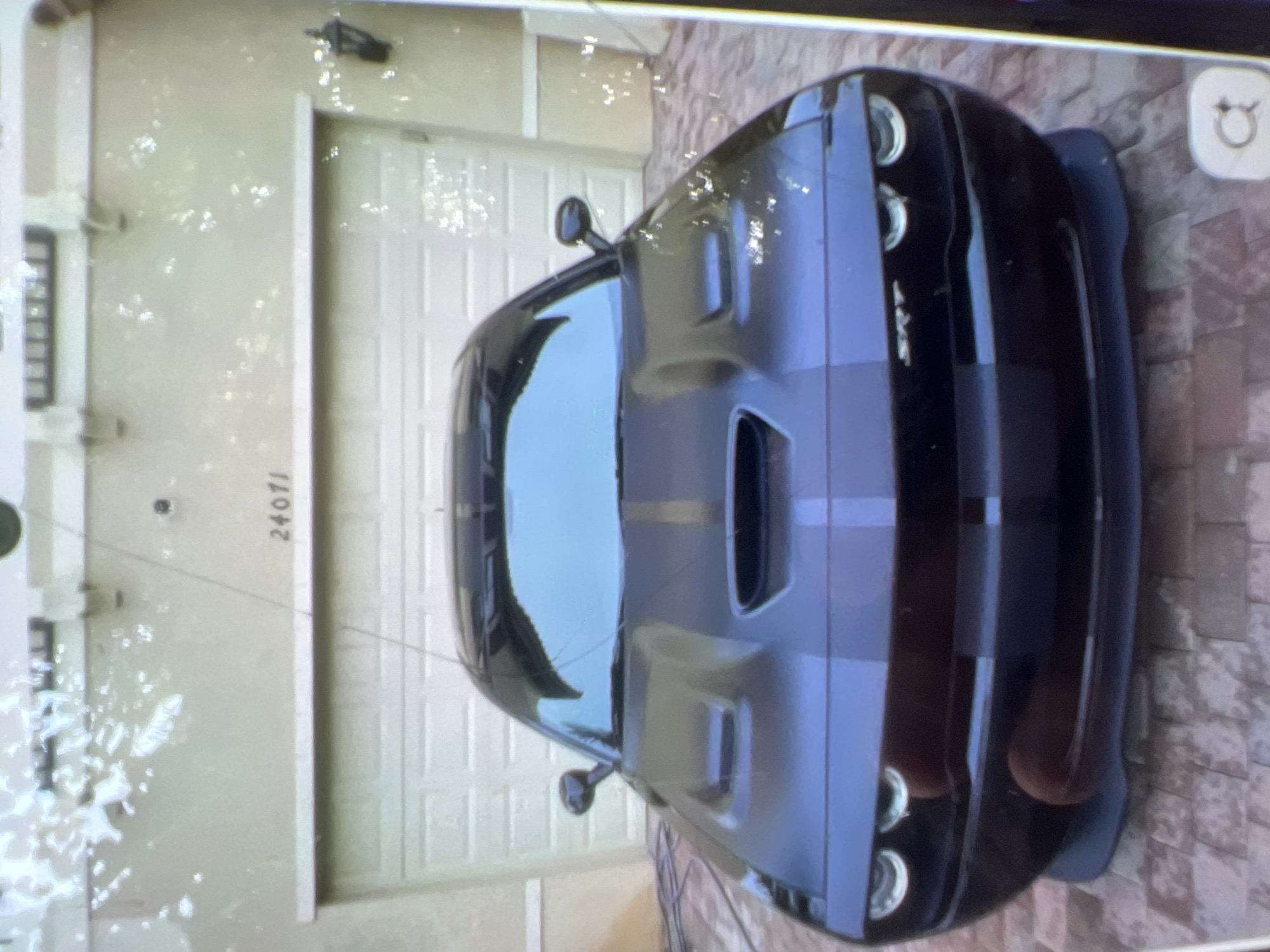 The image shows a black Dodge Challenger SRT parked in front of a garage. The house number "24071" is visible above the garage door. The car has racing stripes and is angled towards the camera, with its headlights and grille clearly visible. The driveway is paved with brick. The scene appears to be a residential area. The image quality is poor, with a grainy texture and some glare. The image is a snapshot, likely taken by a mobile phone. There are no people visible in the image. The lighting suggests it is daytime, with some shadows cast by the car and the house. The background is blurred, indicating a shallow depth of field. The overall impression is of a car enthusiast's pride and joy, showcased in front of their home.