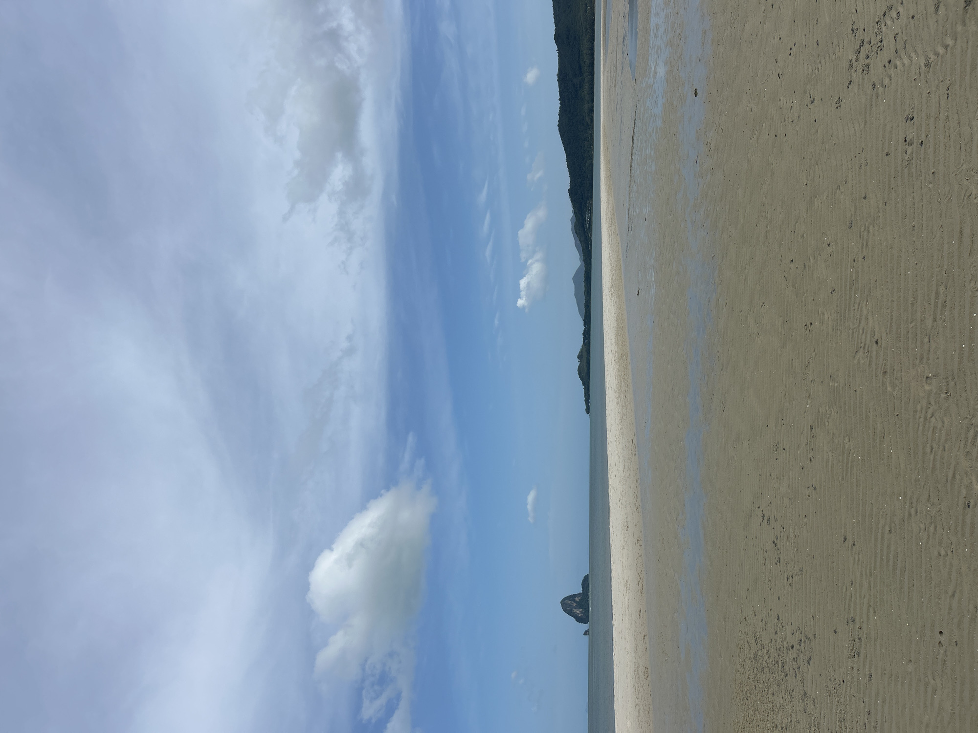 The image depicts a serene coastal landscape in Ko Yao, Thailand. The foreground is dominated by a sandy beach with visible ripples from the receding tide, interspersed with shallow pools of water reflecting the sky. In the mid-ground, the tranquil sea stretches towards the horizon, where a distinctive, solitary rock formation rises from the water to the left. Further in the distance, a chain of lush, green hills forms the coastline. The sky above is a canvas of blue, adorned with scattered white and grey clouds, some of which are wispy and others more voluminous. The overall impression is one of natural beauty and peacefulness, characteristic of a tropical beach setting. There are no people or animals visible in the scene.