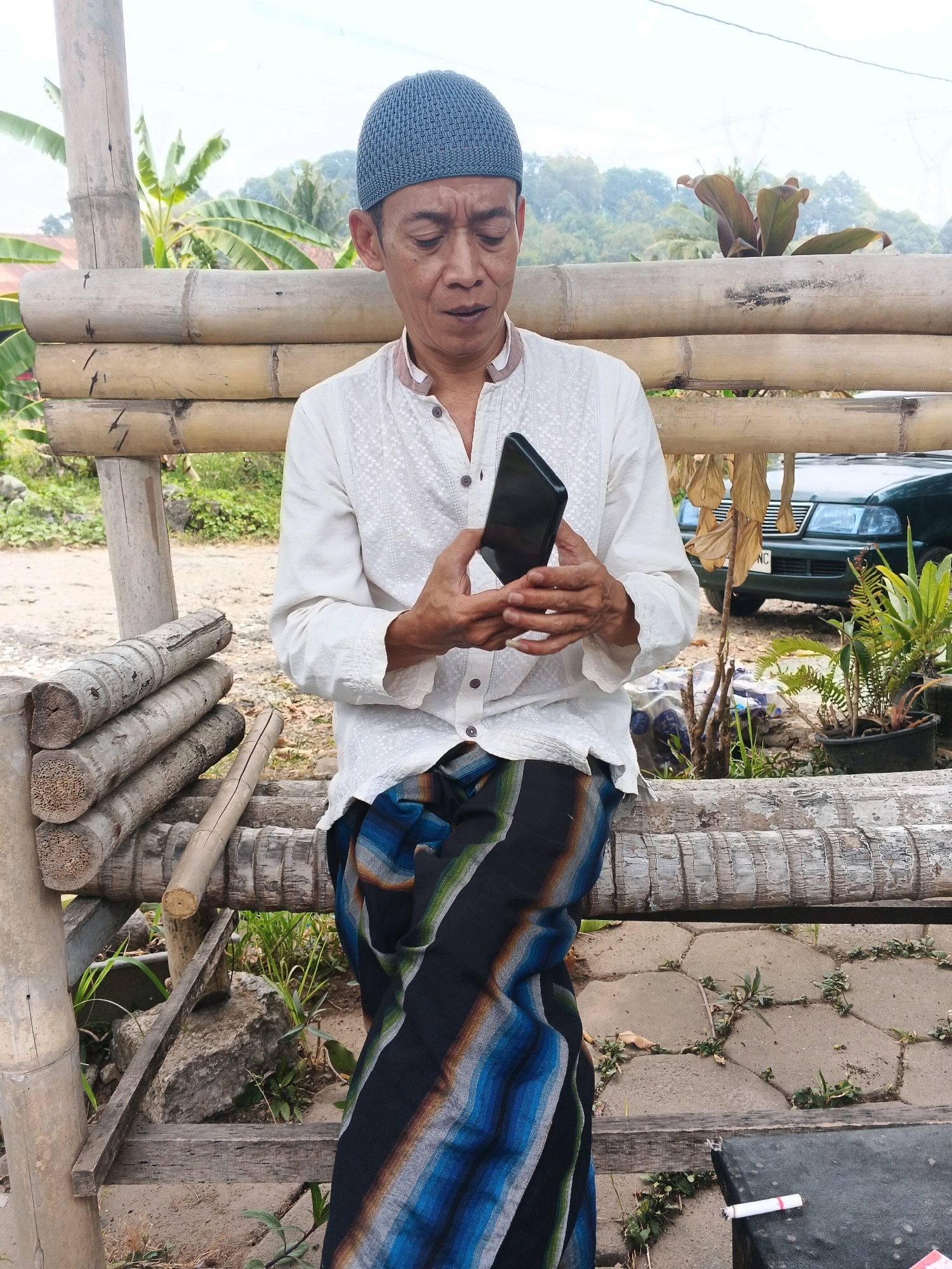 An elderly man wearing a white shirt and a blue skullcap is sitting on a bamboo bench in Ungaran, Indonesia. He is holding a smartphone and looking at it. The man is wearing a sarong with blue, black, and white stripes. In the background, there is a green car and some plants. The bench is made of bamboo poles and has a weathered appearance. There are also some logs stacked on the left side of the bench, also made of bamboo. The ground is covered with stone tiles and some grass. A cigarette butt is visible on a black surface to the right of the bench.