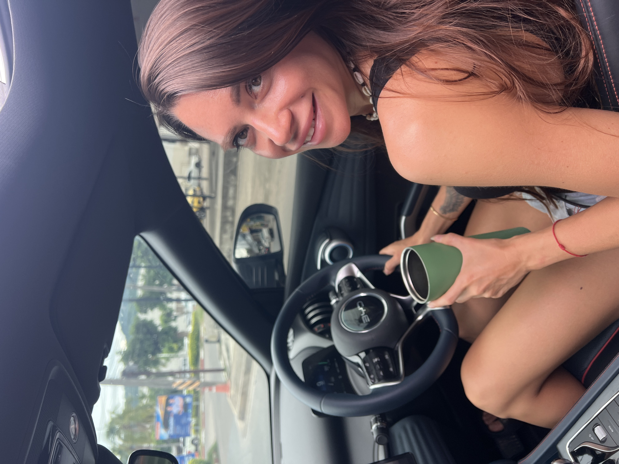 A woman sits in the driver's seat of a car, holding a green tumbler. She is looking over her right shoulder towards the camera with a smile. The car's interior, including the steering wheel with a "BYD" logo, is visible. The woman is wearing a black top and a shell necklace. Her legs are visible, and she is wearing a red string bracelet on her left wrist. Outside the car, a street scene with trees and buildings can be seen, suggesting an urban or suburban environment. The time of day appears to be daytime, possibly late morning or early afternoon, based on the lighting conditions. The location is identified as Baan Bo Phut, Thailand.