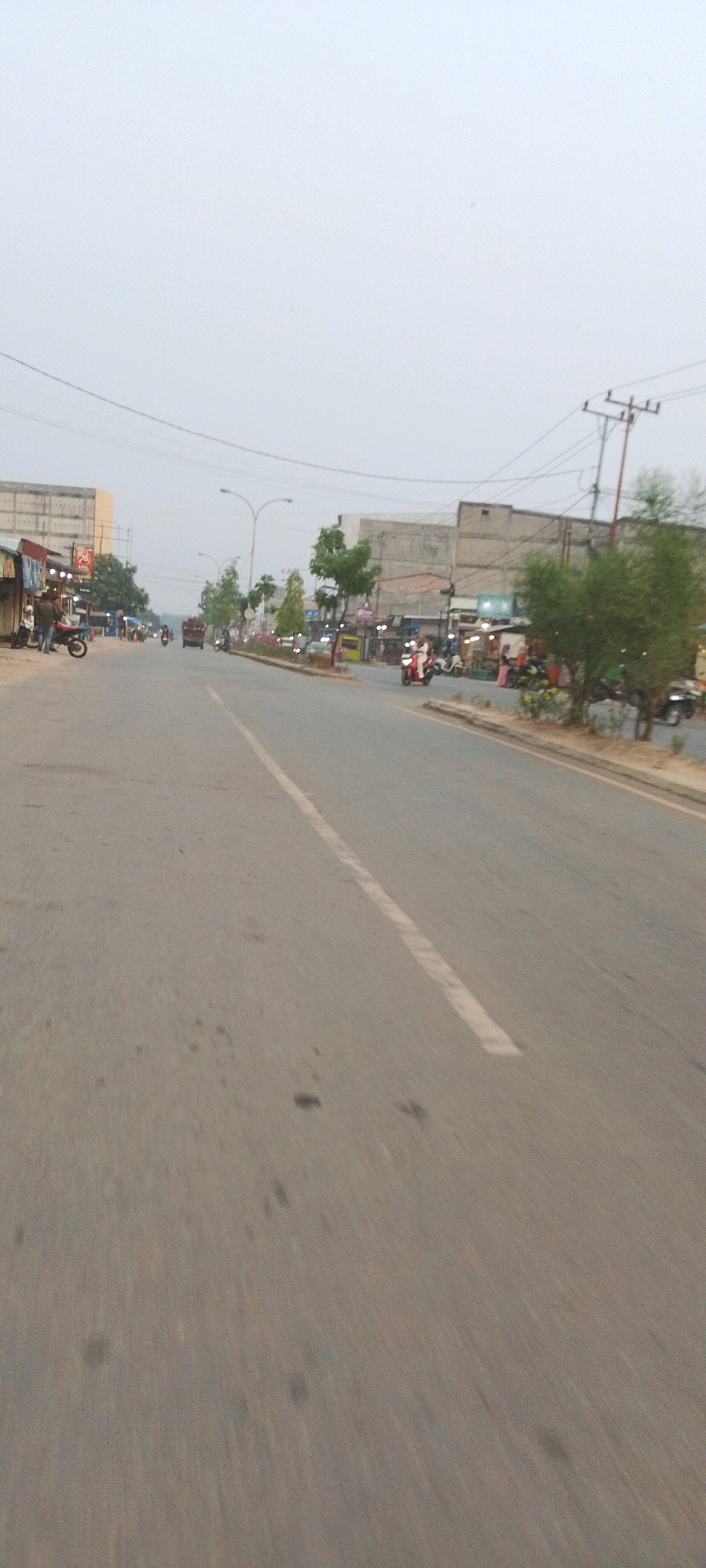 The image depicts a street scene in Lenggadai Hulu, Indonesia, characterized by a two-lane asphalt road with a white dividing line. The sky is overcast, suggesting a dull or hazy day.
On the left side of the road, there are several low-rise buildings, some appearing to be shops or stalls with awnings. A few parked motorcycles and scooters are visible, along with a person standing next to a motorcycle. Further down the road on the left, a small truck with a load is in motion.
The right side of the road features more buildings, also appearing to be commercial establishments, with some of them having outdoor seating or displays. Several motorcycles and scooters are also present on this side, with a few individuals visible. A red scooter is moving along the right lane, and other vehicles are further down the road. Trees and greenery are interspersed along the roadside. Power lines and utility poles are visible in the background, indicating the presence of infrastructure. The overall impression is a bustling street with ongoing commercial activity and vehicular traffic.