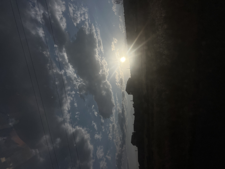 A landscape image features a bright sun shining through a cloudy sky. Power lines run diagonally across the left side. The right side is dominated by dark shadows, obscuring details of the terrain and vegetation. Silhouetted buildings are visible along the horizon line where the sun is positioned. The overall scene is a high-contrast view of a landscape bathed in sunlight.