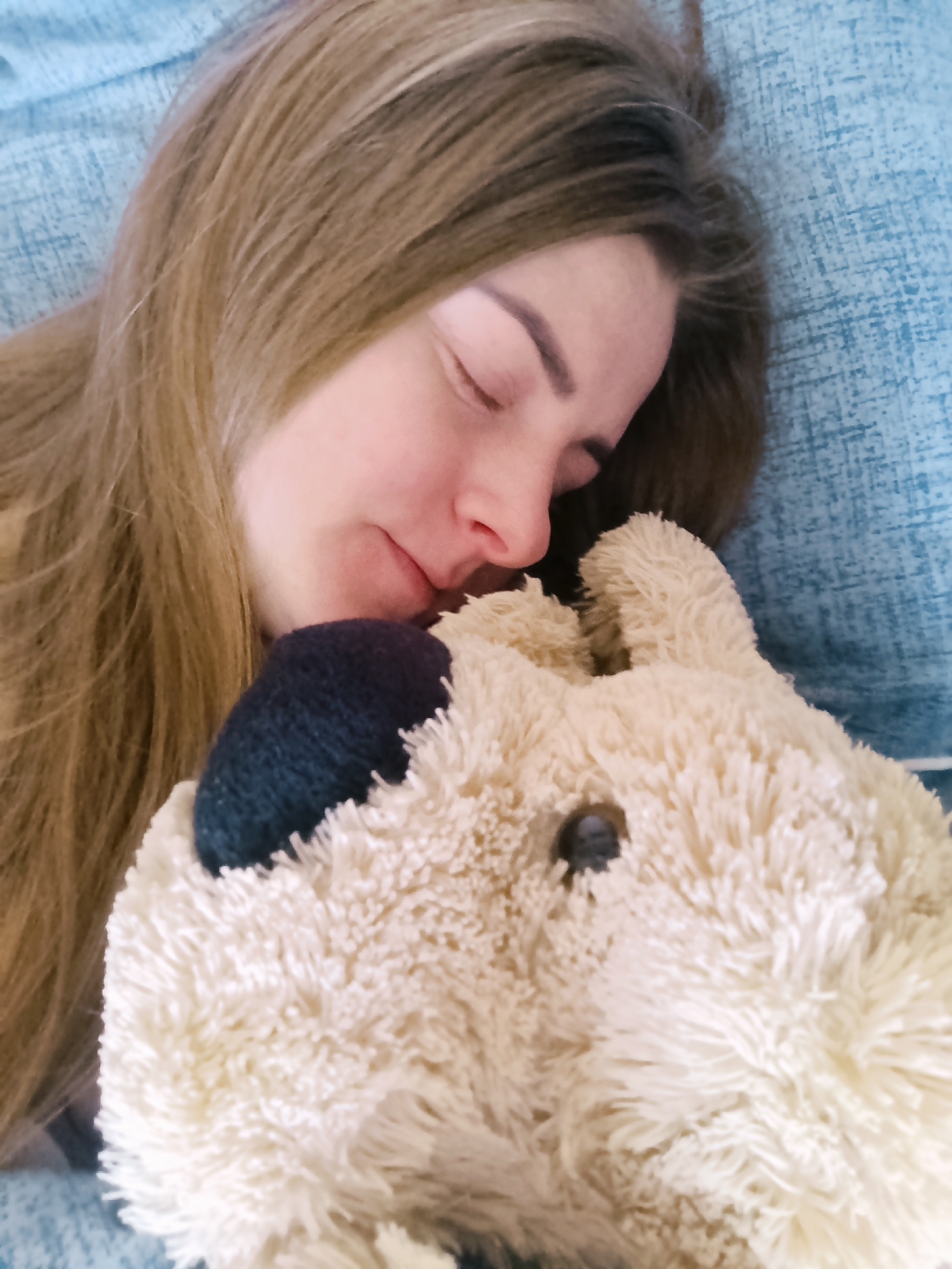 A woman is asleep on a blue couch or bed, with her head resting on a stuffed toy. The woman has long, light brown hair. The stuffed toy is light brown with a dark blue ear. The location is unknown.
