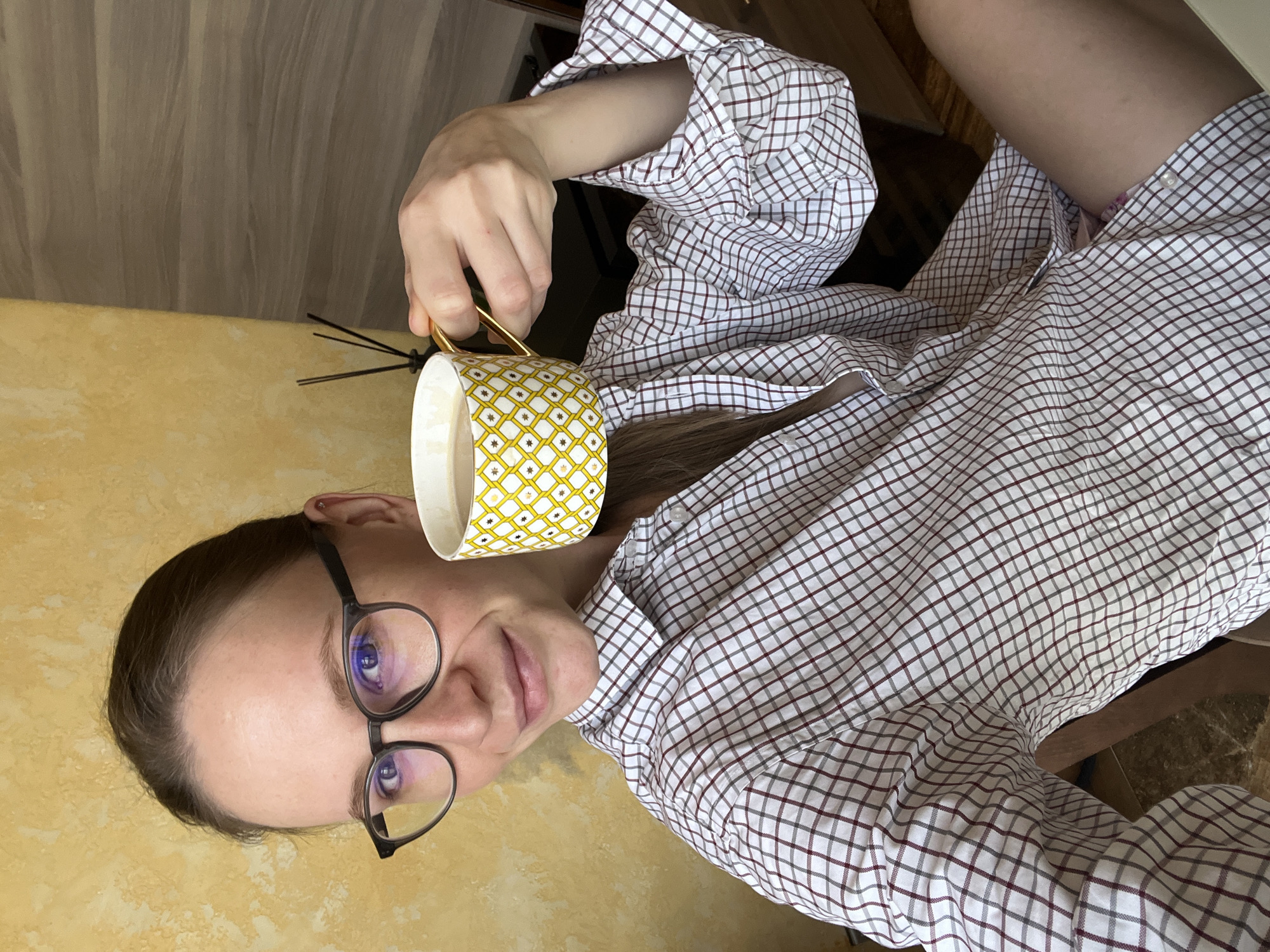 A woman wearing glasses and a white, red, and gray plaid shirt is holding a yellow and white patterned cup with a gold handle. She is sitting and looking directly at the camera. To her left is a yellowish-brown wall with a diffuser. The location is unknown, but it is not possible to verify the location as Khimki, Russia from this image.