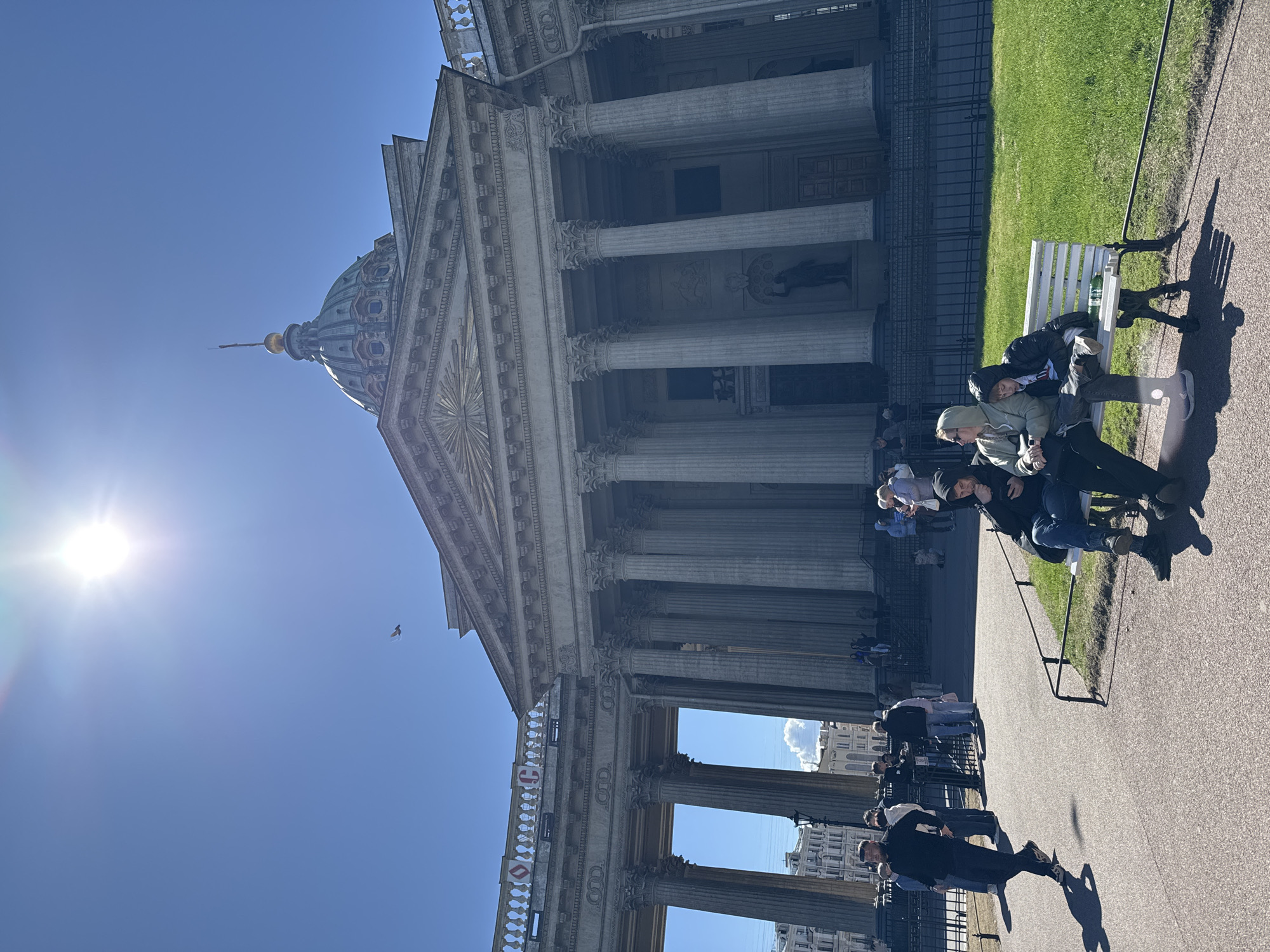The image shows the Kazan Cathedral in Saint Petersburg, Russia, on a bright, sunny day. The cathedral is a large, neoclassical building with many columns and a prominent dome. In the foreground, several people are sitting on benches along a walkway. The walkway is paved and runs parallel to a green grassy area next to the cathedral. The sun is visible in the sky, positioned to the left of the dome. A bird is flying in the sky, near the top of the building.