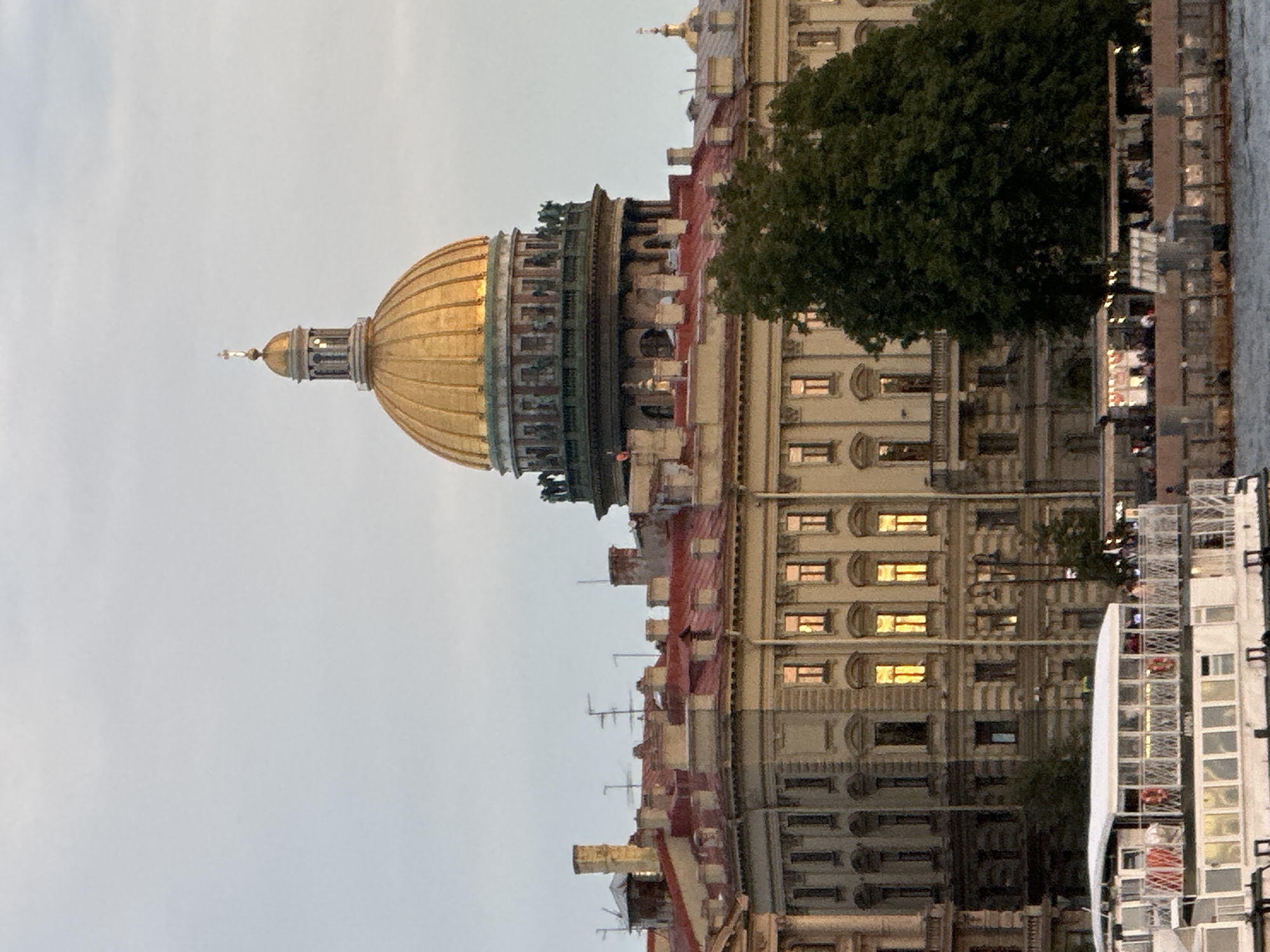 This image captures the grandeur of Saint Isaac's Cathedral in Saint Petersburg, Russia, as viewed from the Neva River. The cathedral's iconic golden dome, topped with a cross, dominates the upper right portion of the frame, reflecting the soft light of an overcast sky. Below the dome, the intricate architecture of the cathedral is visible, with statues adorning its drum.
In the foreground, a white boat with a white roof and railings is docked, likely a tourist vessel, with people visible on its deck. The boat's lower section is white with windows. Further along the embankment, a series of buildings with red roofs and beige facades are visible, interspersed with trees. Some windows in these buildings are illuminated, suggesting indoor activity. A bustling embankment with tables and umbrellas is also discernible, hinting at a lively atmosphere. The overall scene is serene, with the historical architecture of Saint Petersburg set against a cloudy sky.