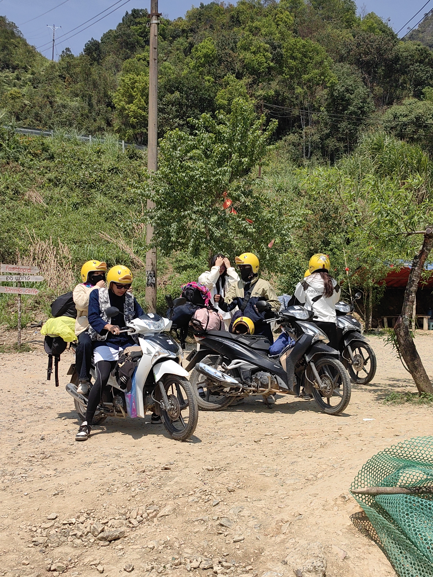 In Phố Bảng Commune, Vietnam, four people in yellow helmets are standing near three parked motorbikes on a dirt road. The people appear to be taking a break, as one is adjusting her helmet. Behind them, a lush green hillside rises. To the right is a tree and a glimpse of a red awning. In the foreground, a green net lies on the ground. A sign post partially visible on the left. Overhead power lines cross the upper portion of the image.
