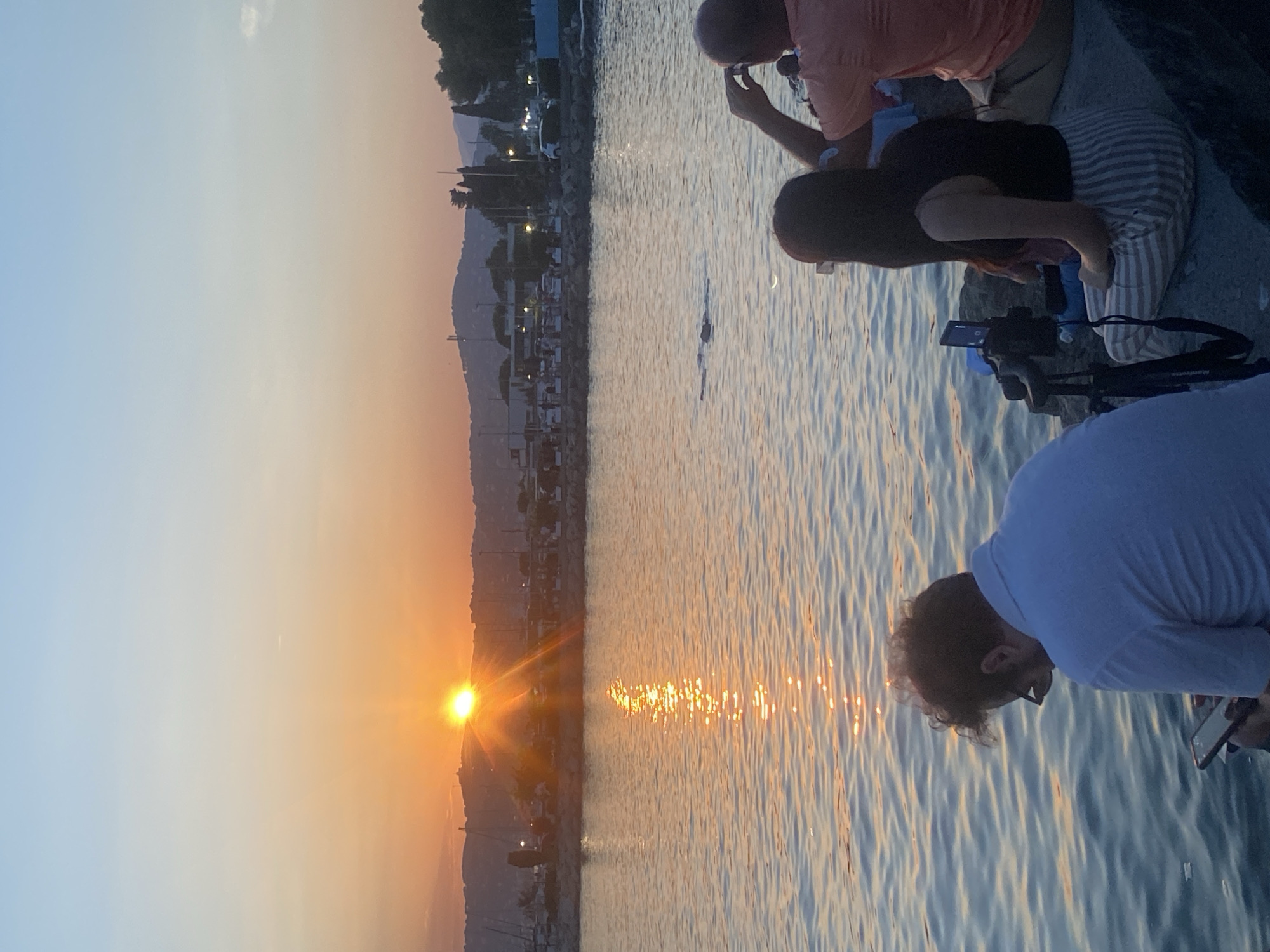 In Antibes, France, at sunset, three people sit on a rocky shoreline facing the water. A man in a light blue polo shirt, wearing glasses, looks down at his phone. Beside him, a woman with long dark hair, wearing a black tank top and striped pants, is seated. A man in an orange shirt is positioned to her right, holding a camera to his face. The sun, a brilliant orange orb, is low on the horizon, casting a shimmering reflection on the calm water. In the background, a marina with boats is visible, backed by a distant hillside dotted with buildings and trees. The sky transitions from a pale blue at the top to warm hues of orange and pink near the horizon. A small boat or debris is visible on the water's surface.