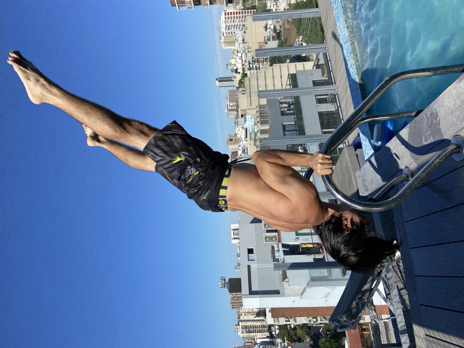 A man in black swim trunks hangs upside down from a metal rail next to a swimming pool. His feet point towards a clear blue sky while his head is closer to the pool deck. The backdrop features a cityscape with various buildings, including apartments and office towers. The pool is partially visible in the upper right corner. The scene is captured in bright daylight.