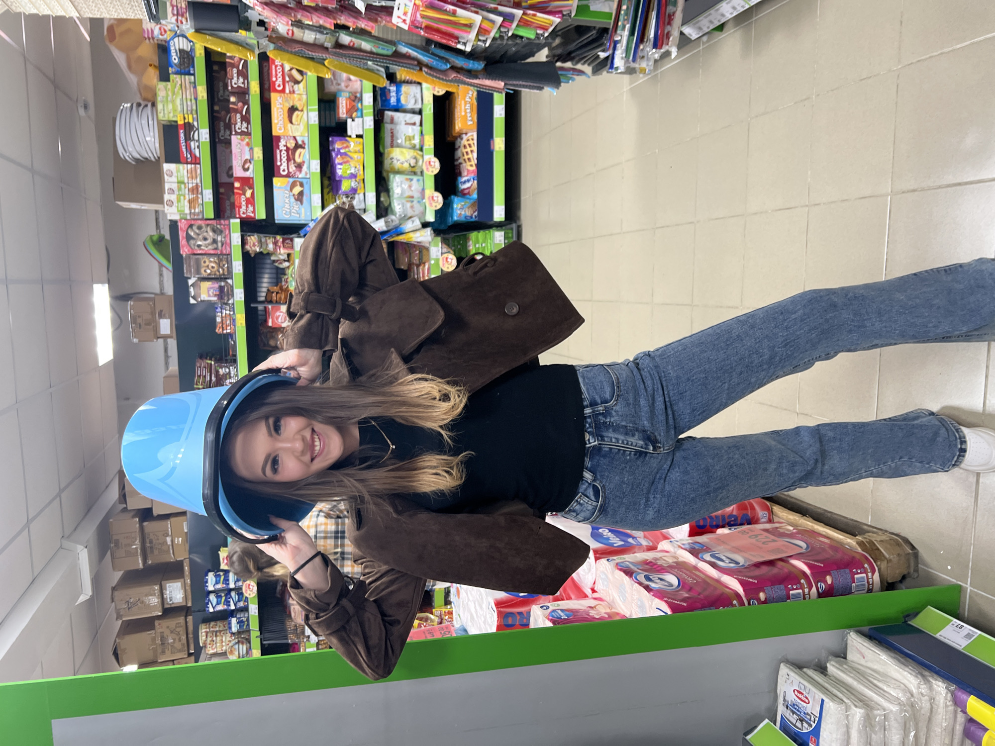 The image depicts a young woman posing inside a supermarket in Chaikovsky, Russia. She has medium-length brown hair, fair skin, and is wearing a black top, a brown suede jacket, and blue jeans. She is holding a bright blue plastic bucket on her head and smiling towards the camera. Shelves stocked with various products, including candy, cookies, paper towels, and cleaning supplies, are visible in the background. The supermarket has a tiled floor and green shelving units.