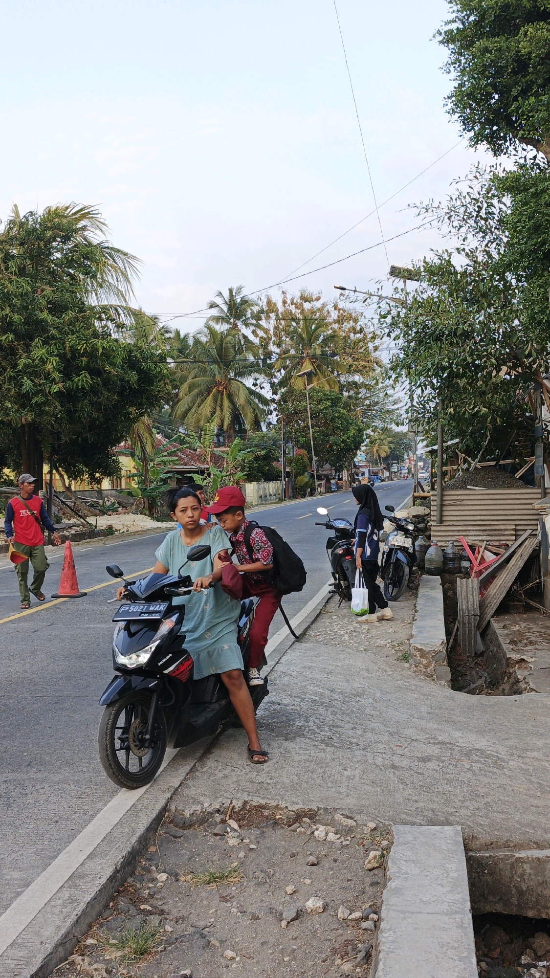 The image depicts a street scene in Cibenda, Indonesia. In the foreground, a woman and a young boy are seated on a black scooter. The woman, wearing a light blue dress, is in the driver's seat, while the boy, dressed in a red shirt, red pants, and a red cap, sits behind her. The boy is carrying a dark backpack and has his arm around the woman. They are both looking forward.
To the left of the scooter, on the asphalt road, a man wearing a red shirt and green pants walks away from the camera. He is carrying a yellow object. An orange traffic cone is visible on the road near him.
To the right of the scooter, on the sidewalk, a young person wearing a black hijab and a dark blue top is standing. They are holding a white plastic bag and appear to be waiting. Behind them, another black scooter is parked.
The environment is tropical, with lush green trees and palm trees lining the street. Buildings and houses are visible in the background. The sky is clear with some scattered clouds. The road appears to be a main thoroughfare, with traffic and activity suggested by the presence of multiple vehicles and people. The sidewalk is made of concrete and shows signs of wear and damage in some areas. The overall impression is that of a typical day in a bustling Indonesian town.