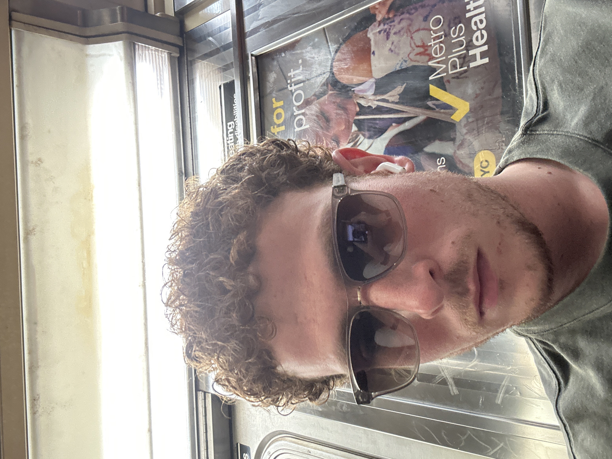 The image displays a young man taking a selfie or being photographed inside a New York City subway car. The photo is oriented ninety degrees counter-clockwise. The man, positioned from the chest up, has curly brown hair and light stubble on his face. He wears dark-rimmed sunglasses, which reflect elements of the interior, and a white earbud is visible in his right ear. He has a neutral expression and is dressed in a grey or olive green top.
The background reveals the interior of the subway. To the viewer's left (if the image were upright), bright rectangular lights illuminate the ceiling panel. To the viewer's right, an advertisement poster is partially visible behind the man. Visible text on the poster includes "for profit.", "eating disabilities", "Metro Plus Health", and "NYC", confirming the New York location. The lighting is bright, suggesting it is daytime.