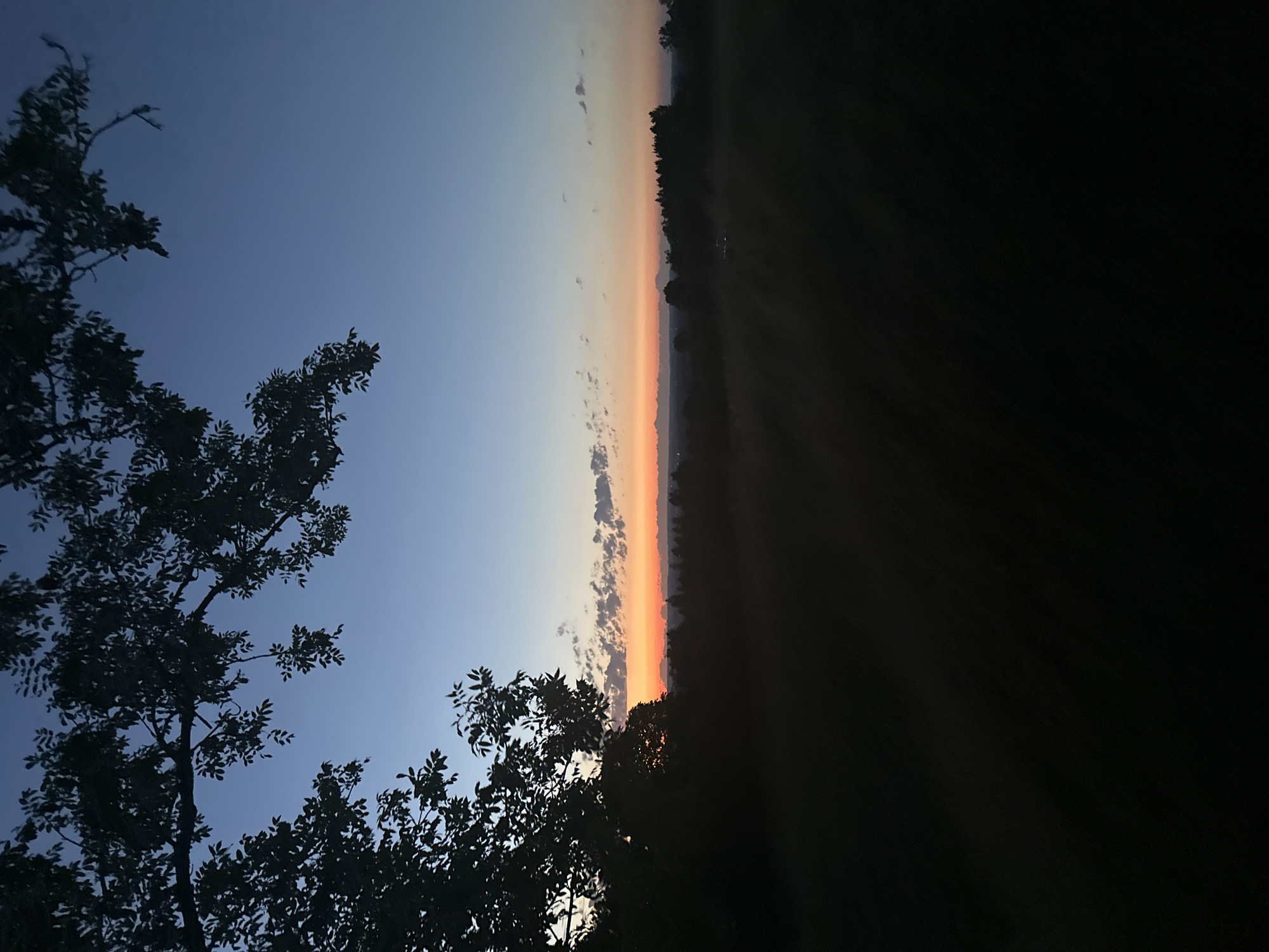 The image depicts a rural landscape at dusk, likely in or near Amerang, Germany. A silhouette of trees and foliage dominates the foreground on the left, framing a view of the horizon. The sky transitions from a deep blue at the top to a gradient of lighter blue and then a warm orange and pink band near the horizon, indicating the setting sun. Scattered clouds are visible in the orange band. The middle ground shows a dark silhouette of a treeline and what appears to be fields or open land, with subtle hints of distant lights. The overall atmosphere is serene and peaceful, characteristic of a rural evening.