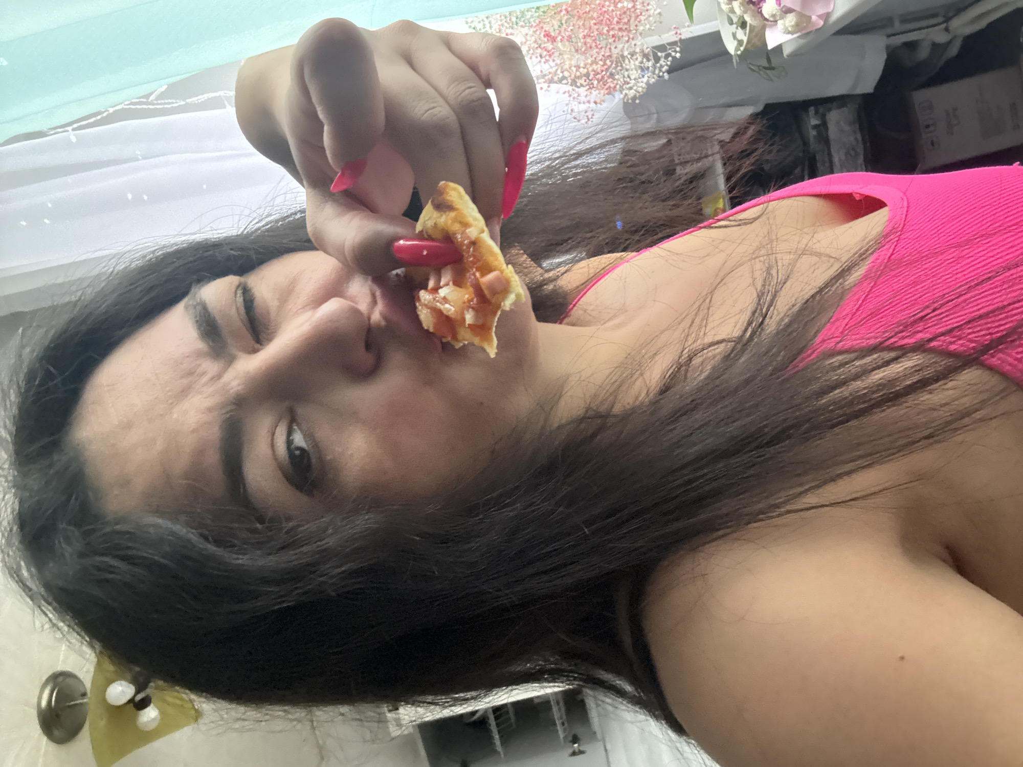 An upward-angled selfie captures a young woman eating a slice of pizza indoors in what appears to be a residential setting in Kauchuk Stantsiya, Russia. The woman, with long, dark hair, is lying down, perhaps on her side or back, looking towards the camera with a playful expression, one eye partially closed as if winking or squinting. She is wearing a bright pink tank top or camisole. Her right hand, adorned with long, bright pink nails, holds a small piece of pizza to her mouth. The pizza appears to have cheese and meat toppings.
The background elements are somewhat blurry but indicate an indoor environment. To the upper left, a light blue or mint green curtain is visible, along with white fabric or another curtain behind her head. Towards the upper right, there is a decorative arrangement of small, delicate pink and white flowers, possibly baby's breath, suggesting a home or personal space. The lighting seems to be natural or from an indoor source, making it appear as if it's daytime. No specific location cues within the image itself indicate Russia, other than the provided context. There is no visible text in the image.