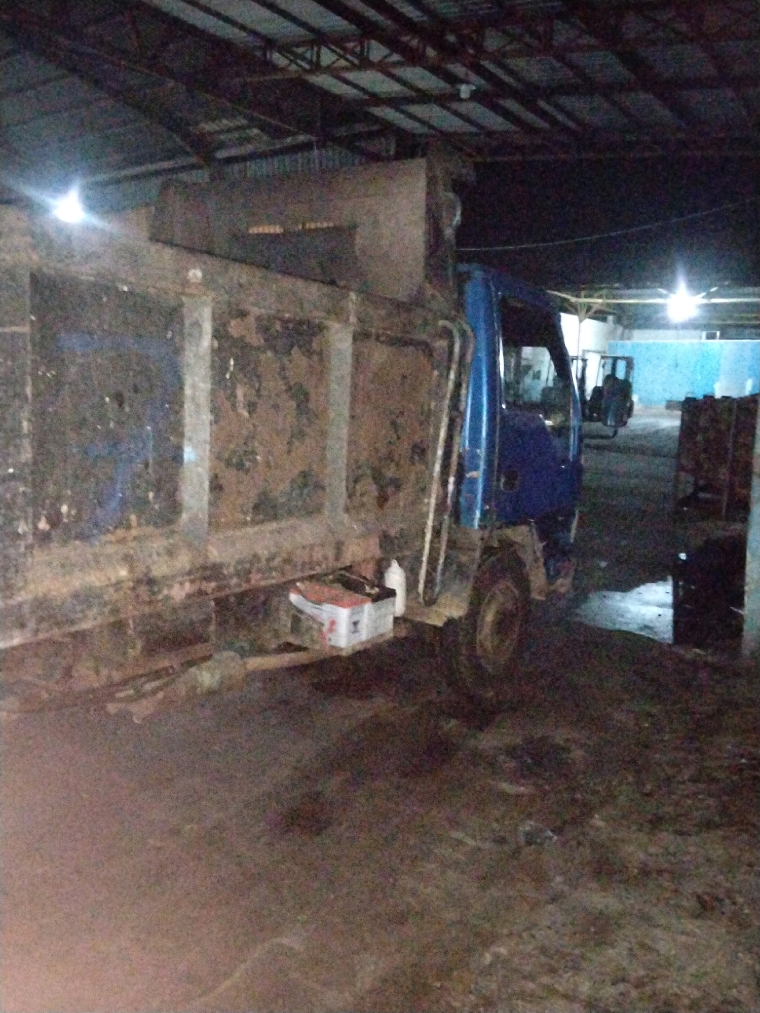 A blue dump truck is parked inside a dimly lit warehouse in Moro, Indonesia. The truck's bed is tilted and appears to be carrying a load of dirt or debris, with visible rust and dirt on its exterior. A small white box with red markings is placed under the truck's bed. In the background, other vehicles and structures are partially visible, including what appears to be another truck or vehicle and a blue wall. The floor of the warehouse is wet and dirty. The lighting suggests it is nighttime or the warehouse is poorly illuminated.