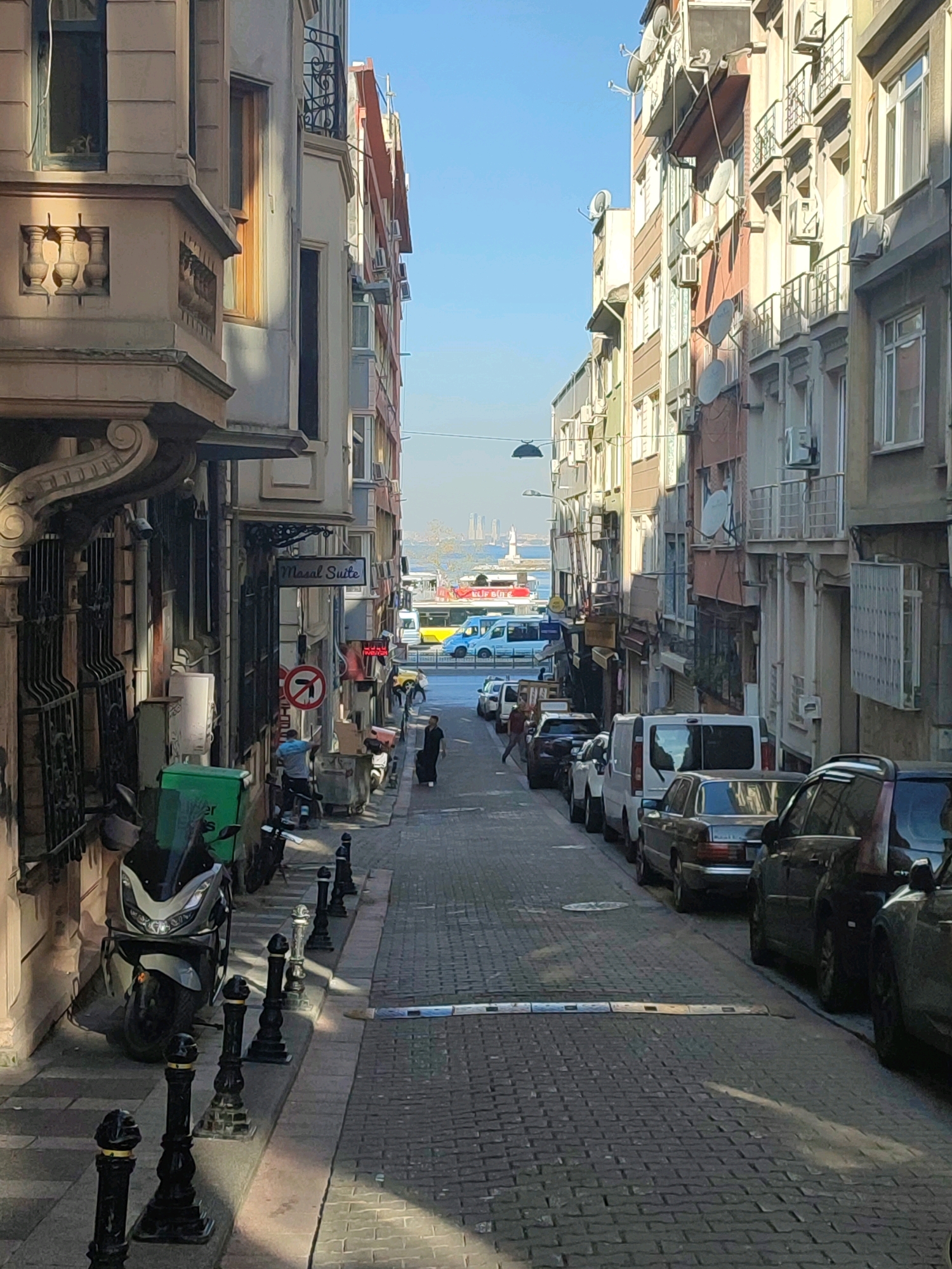 The image depicts a narrow, cobblestone street in Kadıköy, Turkey, likely a residential or commercial area. The street is lined on both sides by multi-story buildings with a mix of architectural styles, many featuring balconies and air conditioning units. The buildings are painted in various muted colors. On the left, a motorcycle is parked near a green delivery box. Further down the street, several vehicles are parked, including vans and cars. In the distance, the Bosphorus Strait is visible with a lighthouse or tower on the horizon. The sky is clear and blue. There are a few individuals present, including one person walking down the street and another near a dumpster. The scene appears to be captured during daylight hours.