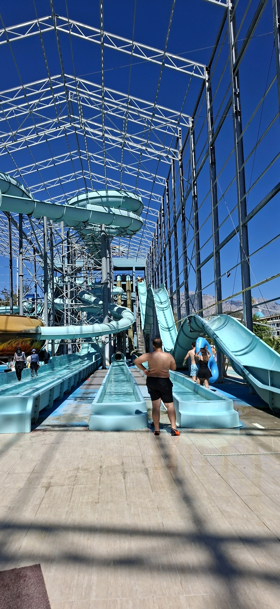 The image shows an indoor water park in Kemer, Turkey, under a large, white, geodesic dome-like structure with a metal framework, revealing a clear blue sky. The water park features several bright blue water slides of various heights and configurations, some of which are enclosed tubes. In the foreground, a man with dark hair, wearing black shorts, stands with his hands on his hips, facing away from the camera towards the water slides. To his left, two individuals, one wearing a patterned headscarf and a dark top and trousers, the other with a similar headscarf and dark trousers, walk away from the camera on a wet surface. To the right of the man, a woman in a black dress and a man in blue shorts are walking towards a blue inner tube. The ground appears to be tiled and wet in some areas. The overall scene depicts a lively water park environment.