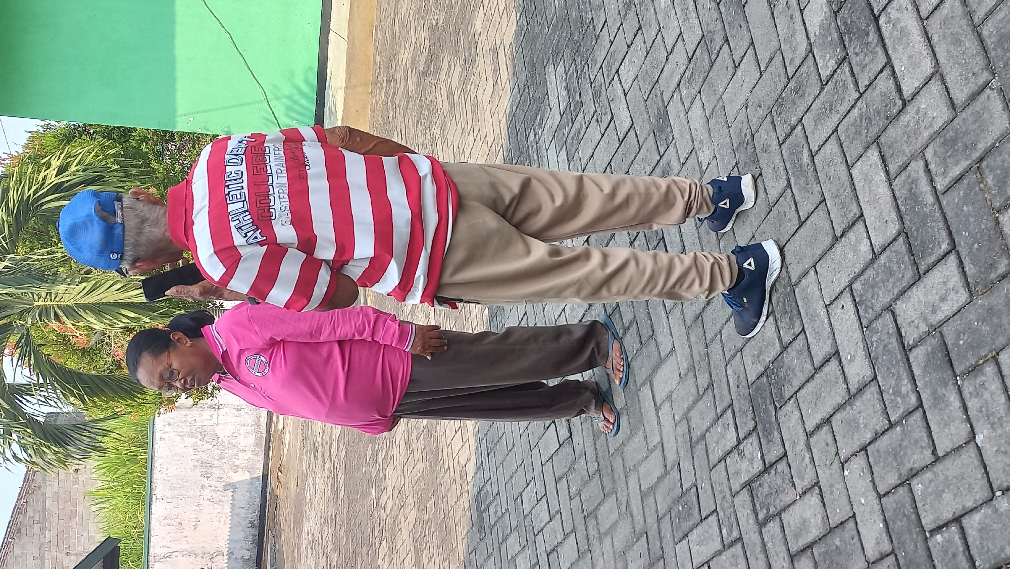 In Kayen Kidul, Indonesia, two individuals stand on a paved outdoor area. On the left, a woman with glasses and dark hair pulled back wears a pink polo shirt and brown pants, with blue flip-flops on her feet. To her right, a man with a blue baseball cap and gray hair is seen from behind, wearing a red and white striped polo shirt and tan pants, with dark blue athletic shoes on his feet. He holds a black mobile phone in his right hand, appearing to take a photo or video. Behind them, a palm tree and green wall are visible. The ground is covered in grey brick pavers.