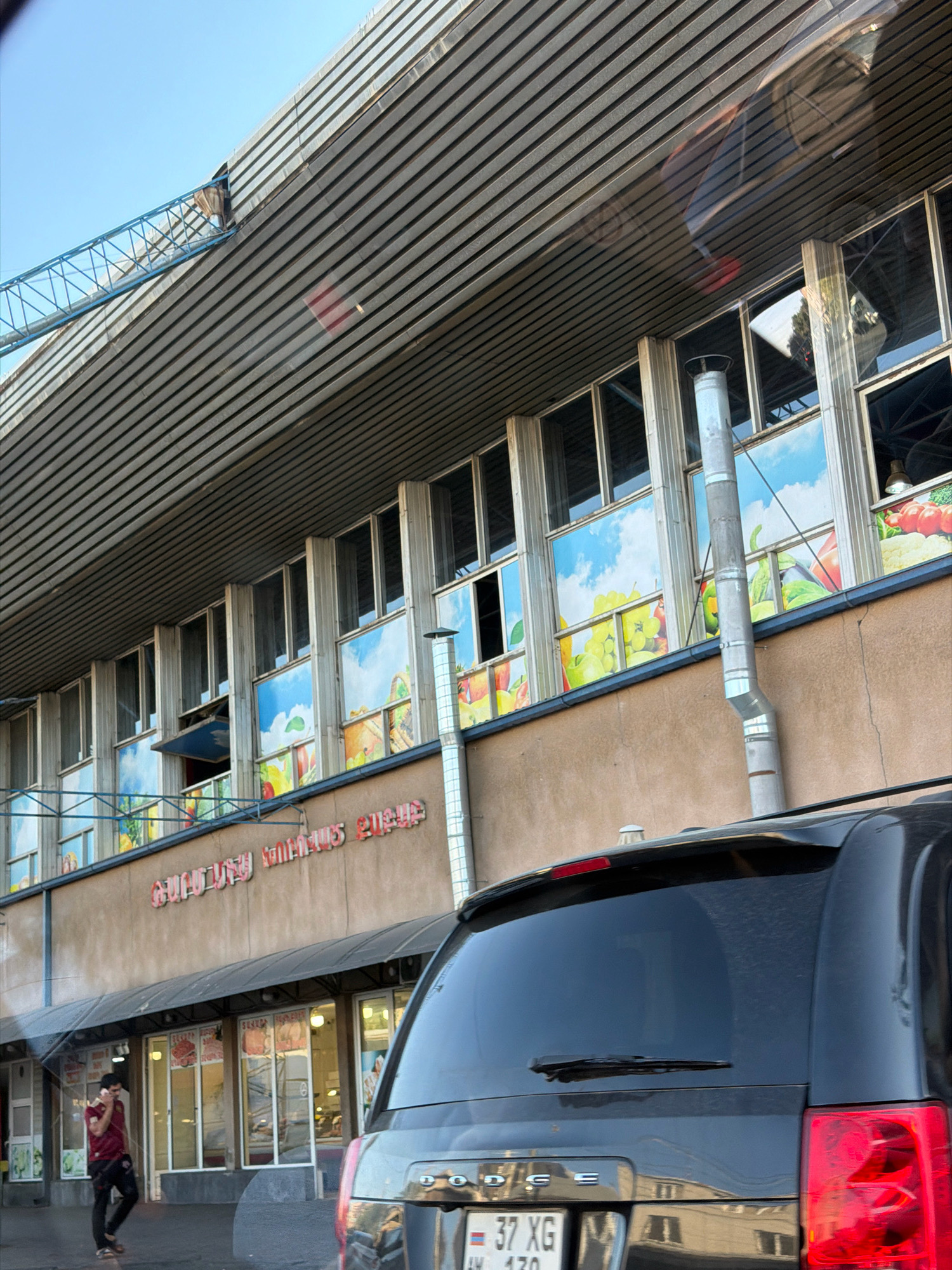 The image shows a market building in Yerevan, Armenia, with large windows adorned with fruit and vegetable graphics. The building has a distinctive ribbed metal roof and a beige-colored facade. A black minivan is parked in front of the building, and a man in a red shirt and black pants is walking and talking on his phone. There is a blue construction crane visible in the upper left corner of the image. The overall scene suggests a busy urban environment with commercial activity.