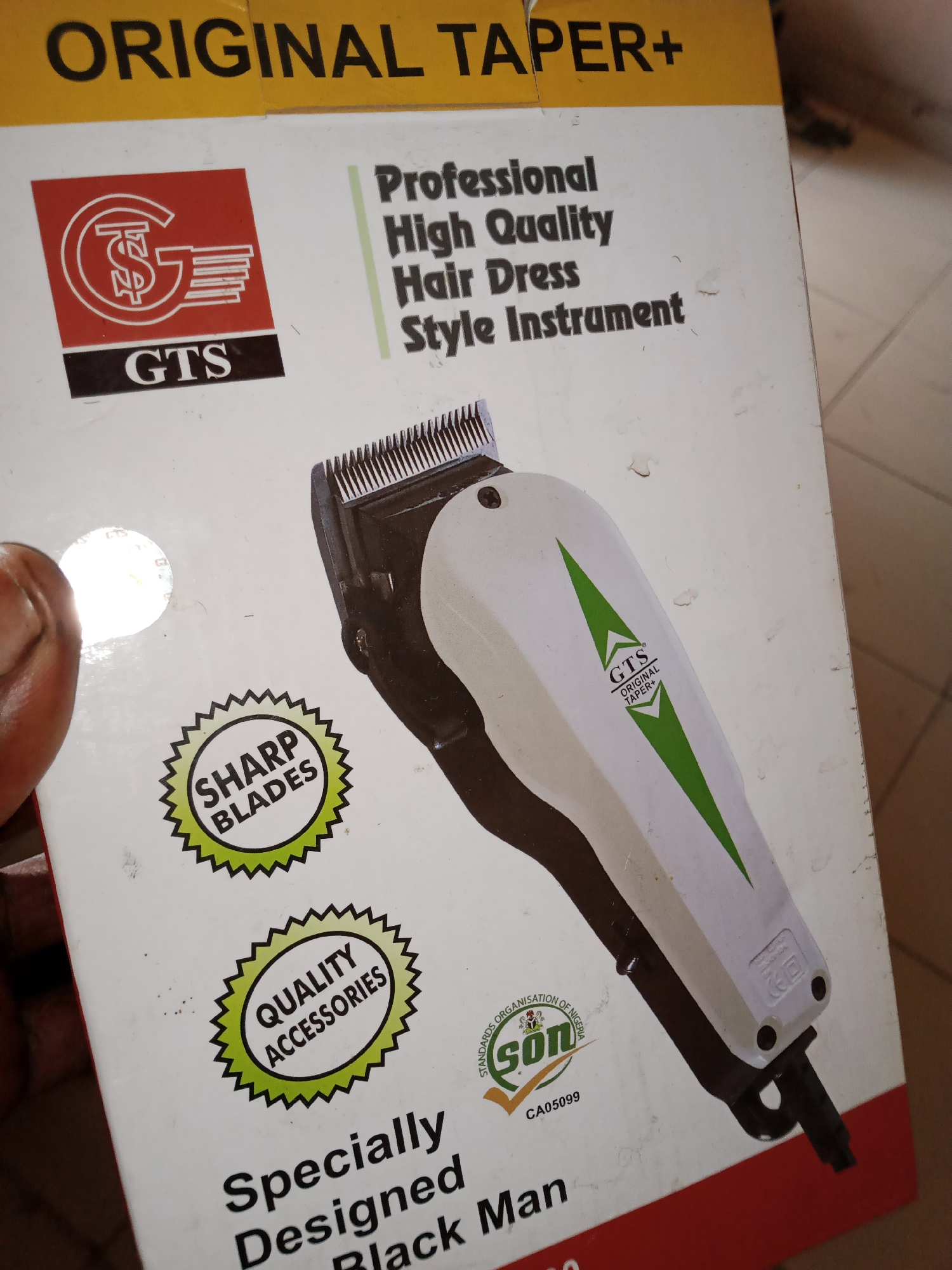 The image shows the front of a product box for a "GTS Original Taper+ Professional High Quality Hair Dress Style Instrument". The box is primarily white with red and yellow accents. A large portion of the box features a diagram of a hair clipper, predominantly white with dark brown and green accents. There are also several circular seals with text: one that says "SHARP BLADES" and another that says "QUALITY ACCESSORIES". A third seal shows "SON CA05099" and "STANDARDS ORGANISATION OF NIGERIA". The text "ORIGINAL TAPER+" is prominently displayed at the top in yellow lettering. There is no indication of a specific location within Jalingo, Nigeria.