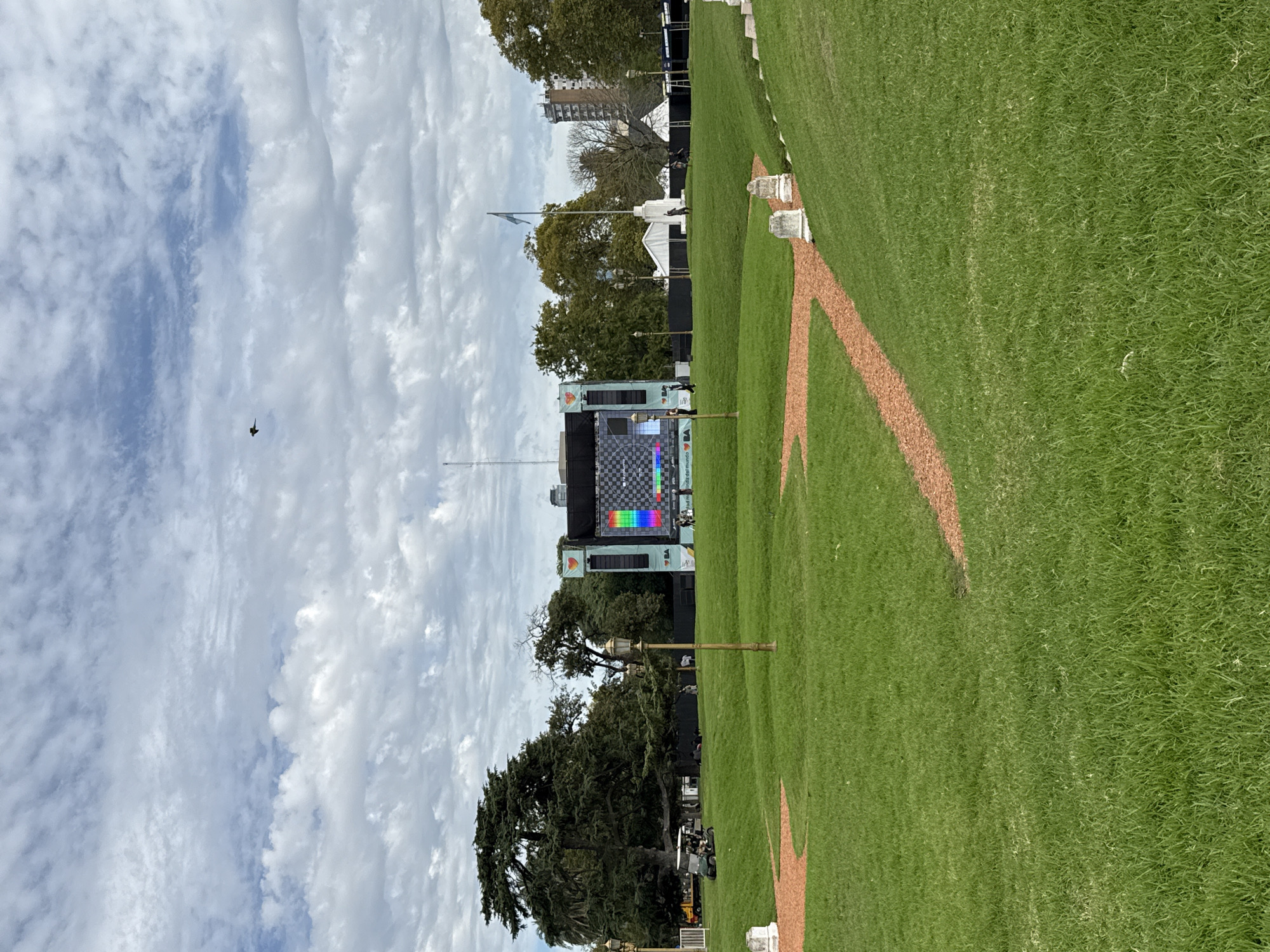 A landscape image displays a green, well-maintained field adjacent to a structure resembling an information display tower or screen, set against a backdrop of sky with scattered clouds. The field is delineated by a light brown, dirt-like material forming a path along its border. The information display tower features a checkerboard pattern and a rainbow-colored display towards the center, topped with a logo or symbol. The structure and field are partially surrounded by trees and urban buildings are visible in the background. A flag is seen next to a white building. The image is shot in Buenos Aires, Argentina. A bird flies in the sky.