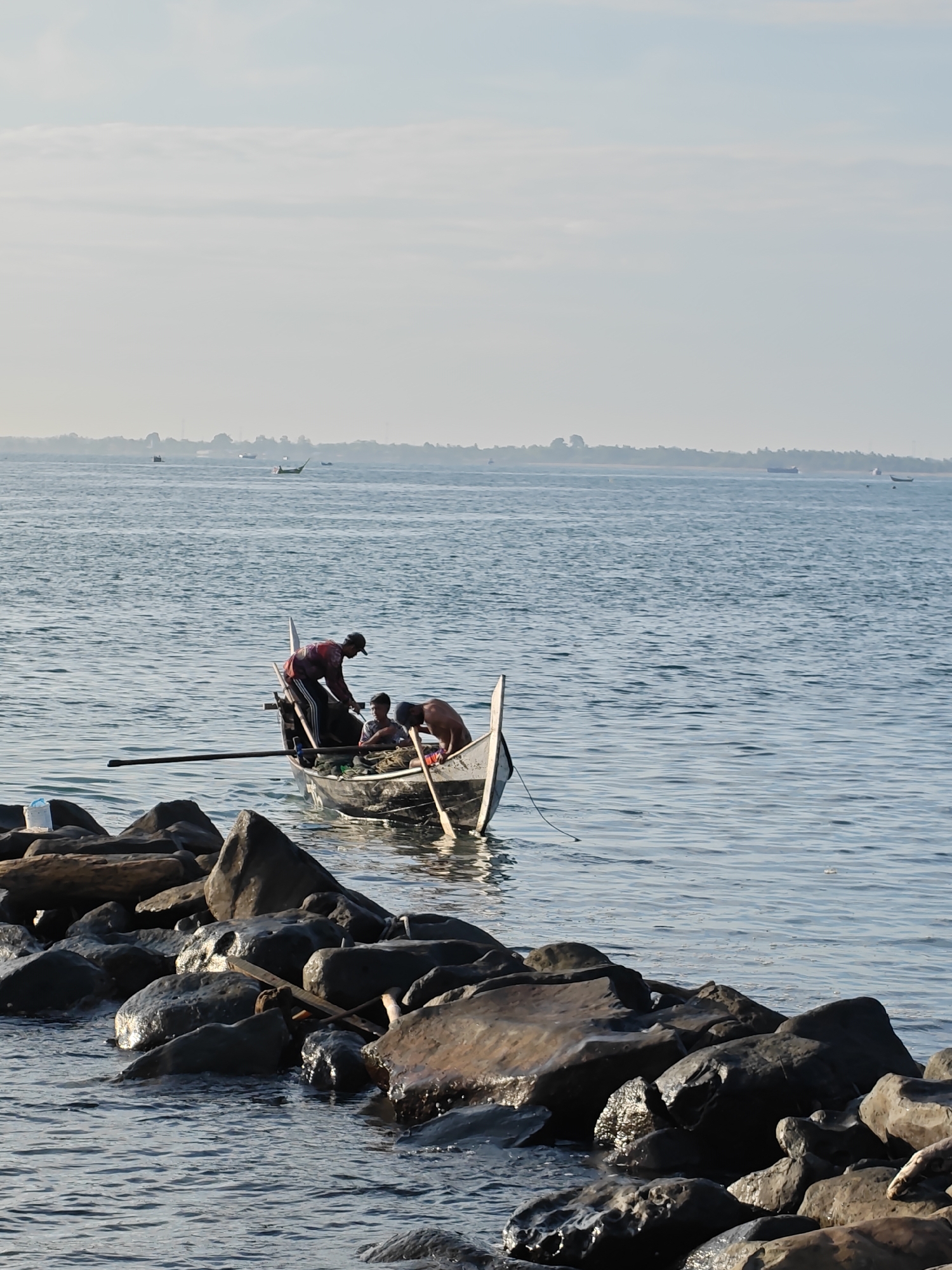 The image depicts three individuals aboard a small, weathered boat close to a rocky shoreline. The boat appears to be a fishing vessel, equipped with oars. One man in a red shirt stands bent over near the stern of the boat, while another leans over the side. A third person sits in the boat's interior. The boat is situated near a collection of dark, wet rocks that form part of a coastal defense. In the background, the calm sea stretches to the horizon under a pale sky. Other boats are visible in the distance. Based on visual clues, including architectural styles and the boat design, the location is Lhokseumawe, Indonesia.