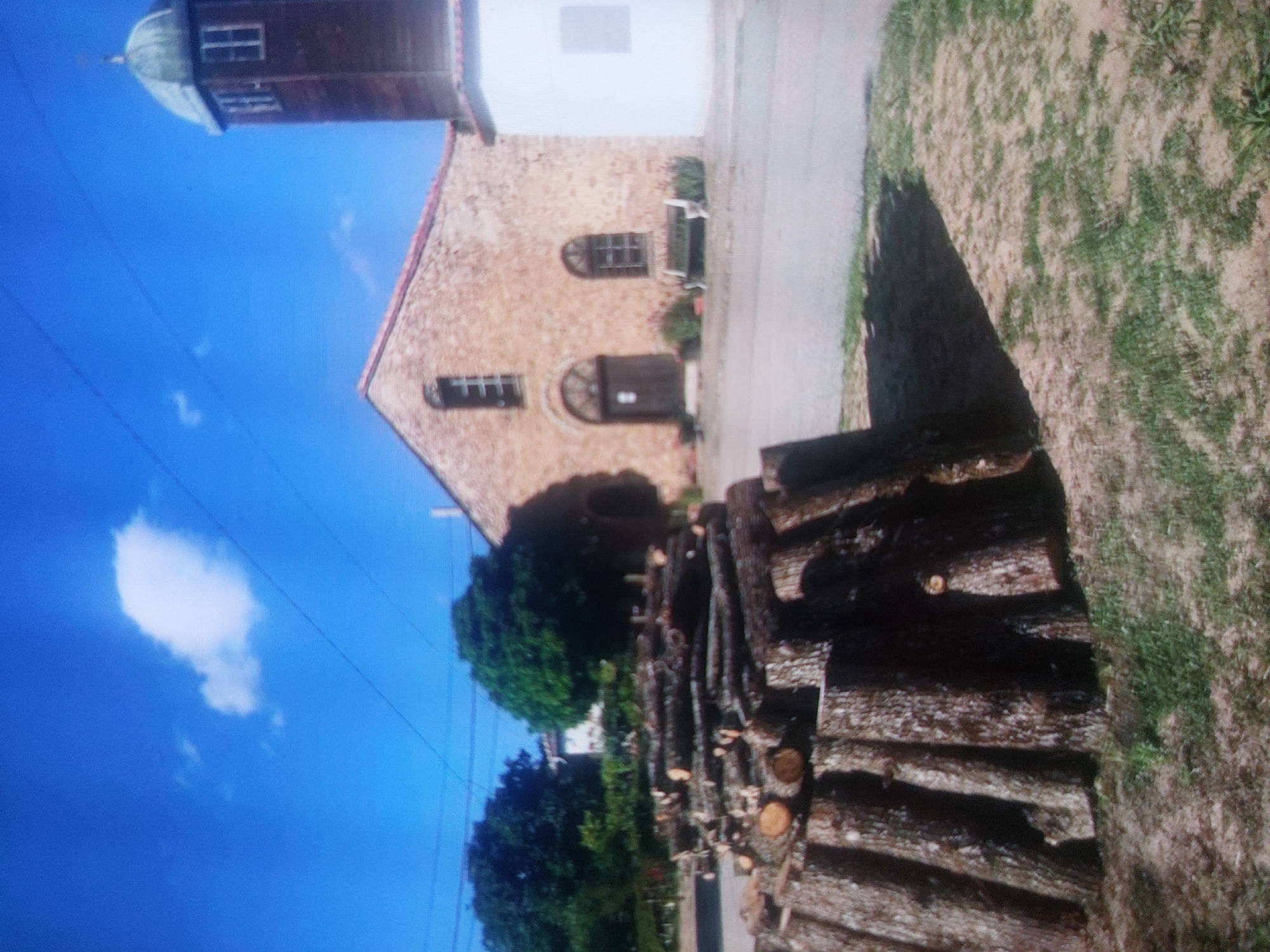 A ground-level, landscape-oriented image displays a scene in Tsarevo, Bulgaria.
The main focal point is a tan building with an arched doorway and windows. Attached to the building's left is a taller brown tower with windows and a dome on top. The sky is blue with a few white clouds in the upper left.
In the right foreground, there is a pile of logs. To the right of the logs, there's grassy terrain. Trees are observed adjacent to the building and in the background. Utility wires are also visible, stretching across the sky.
