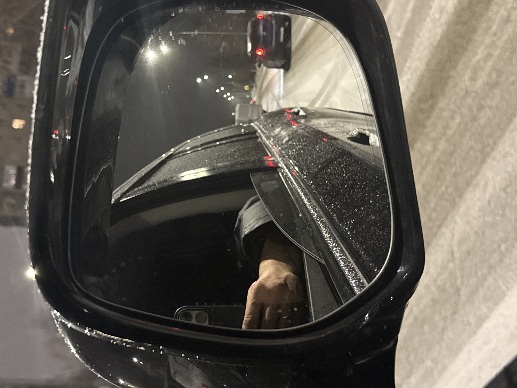 The image shows a side mirror of a vehicle, reflecting a nighttime scene. The mirror's surface is wet with water droplets. Reflected in the mirror is the car's roof, an arm and hand holding a smartphone, and a road with streetlights. The vehicle behind is emitting red light. There are buildings to the side, one of which is light colored. The mirror itself is black and reflecting light in various spots. The location is Nizhnevartovsk, Russia.