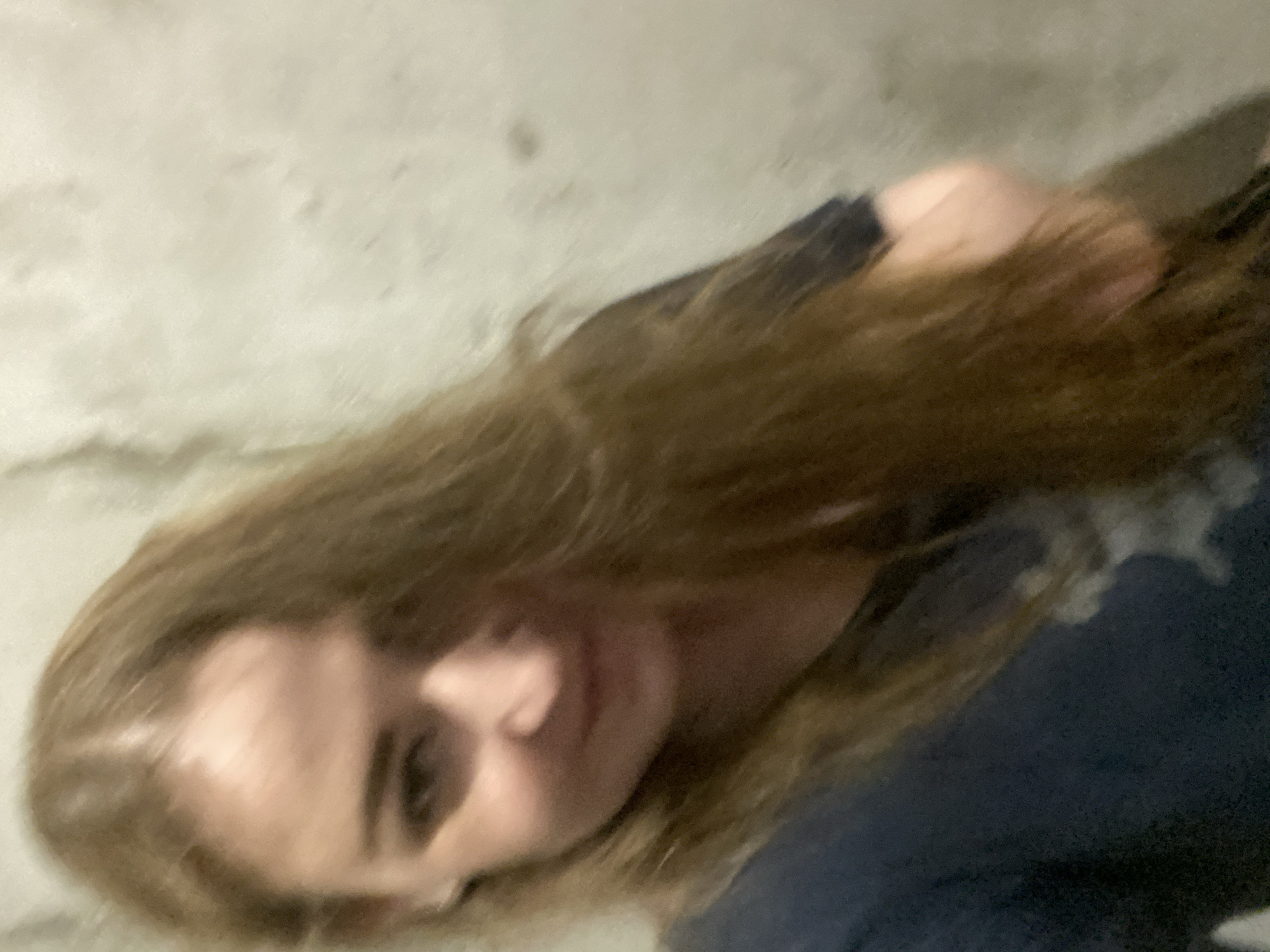 A low-angle, blurry, close-up, indoor selfie shows a young woman with long, light brown hair that falls over her face. She is wearing a dark blue t-shirt. The background is a neutral-colored, textured wall. The lighting is dim and diffused, casting soft shadows. The woman appears to be looking slightly to her left.